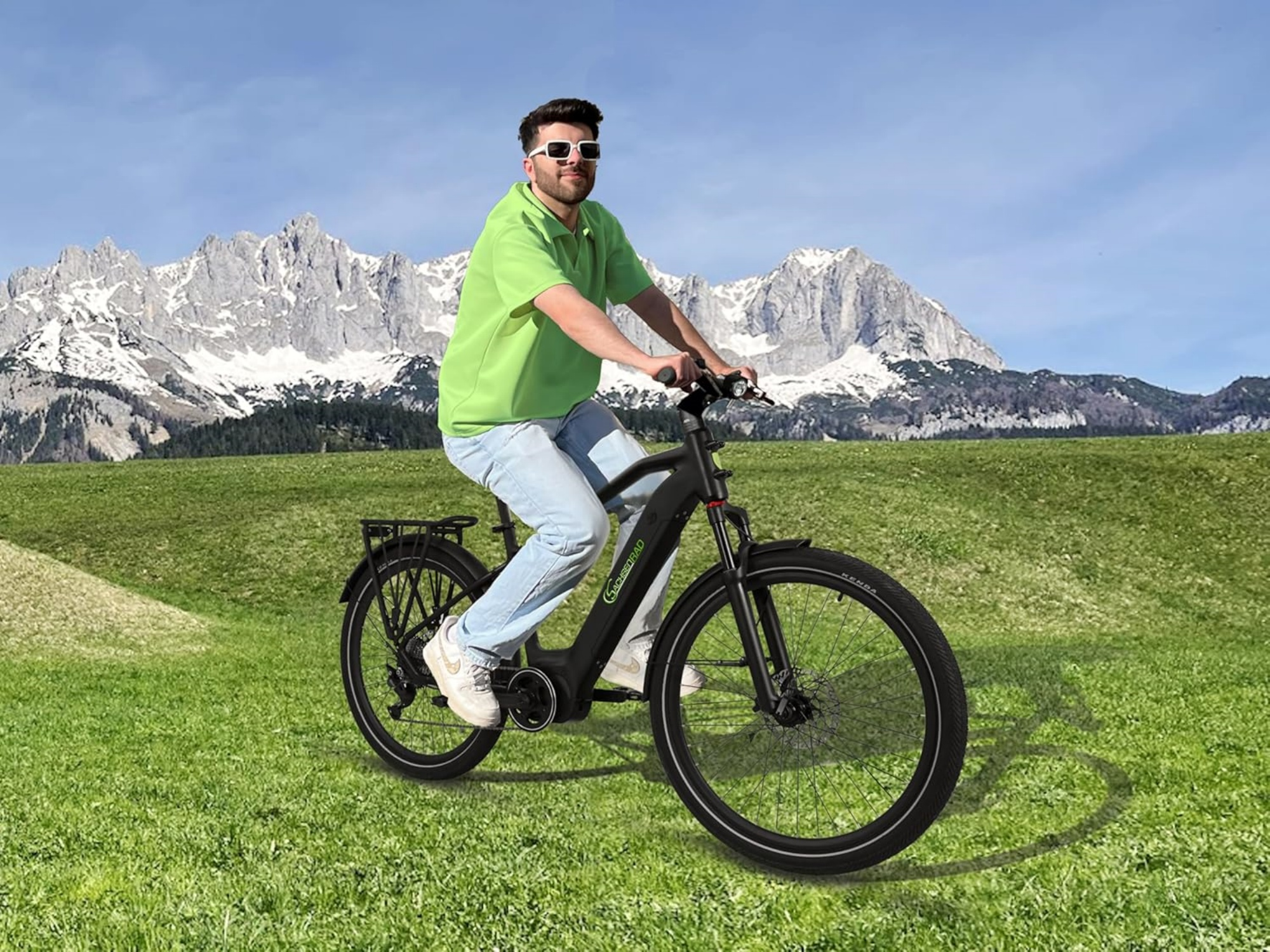 Mann fährt mit einem schwarzen Fahrrad auf Gras, mit Bergen im Hintergrund. Blauer Himmel und weiße Wolken.