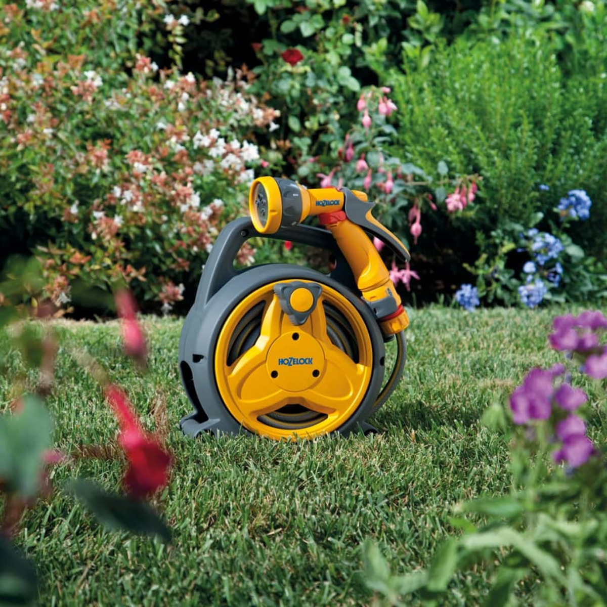 Gelb-graue Gartenschlauch-Trommel auf grünem Gras, umgeben von Blumen und Pflanzen.