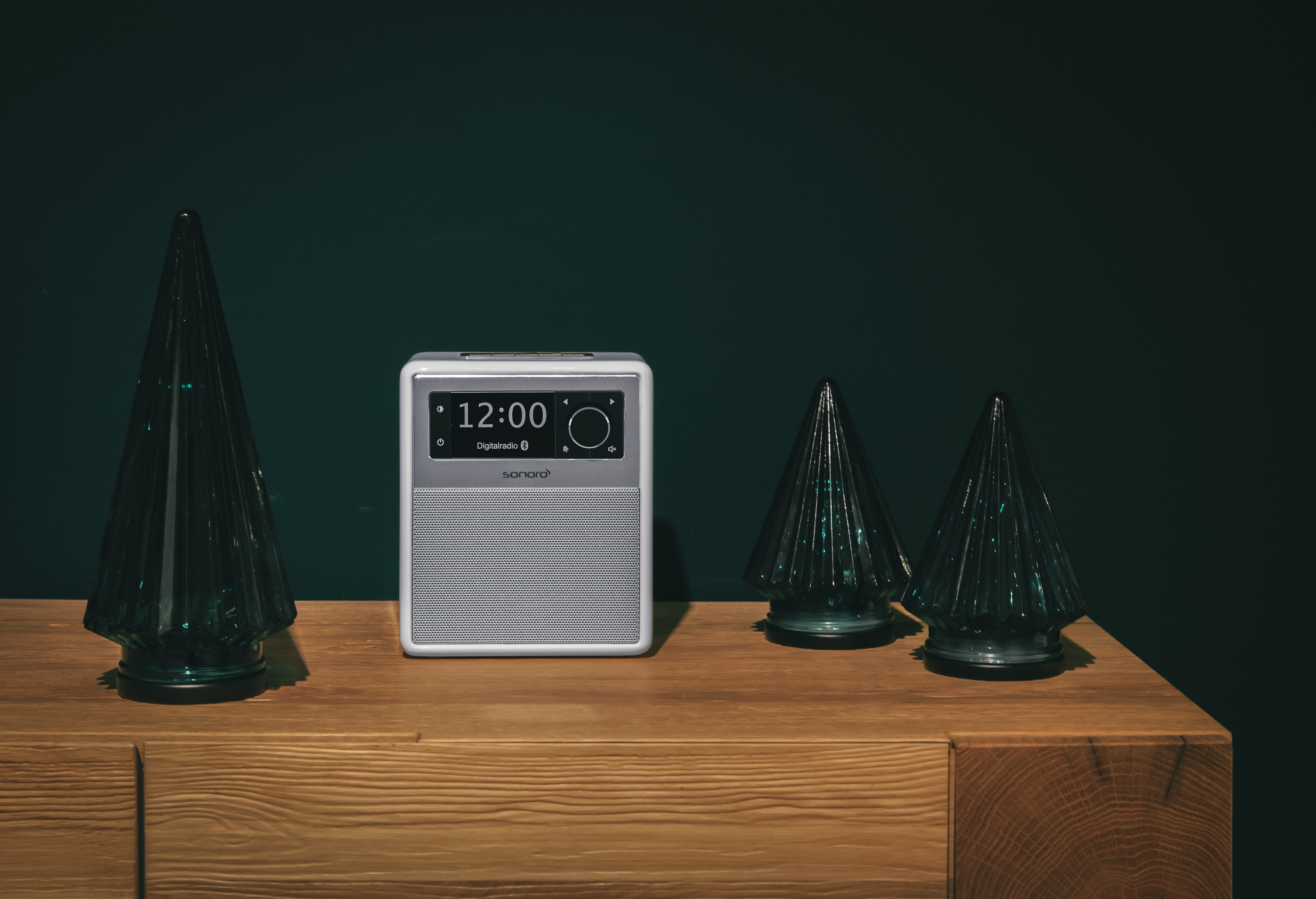 Ein weißes und silbernes Digitalradio mit Bildschirm und drei grünen dekorativen Glastannen.