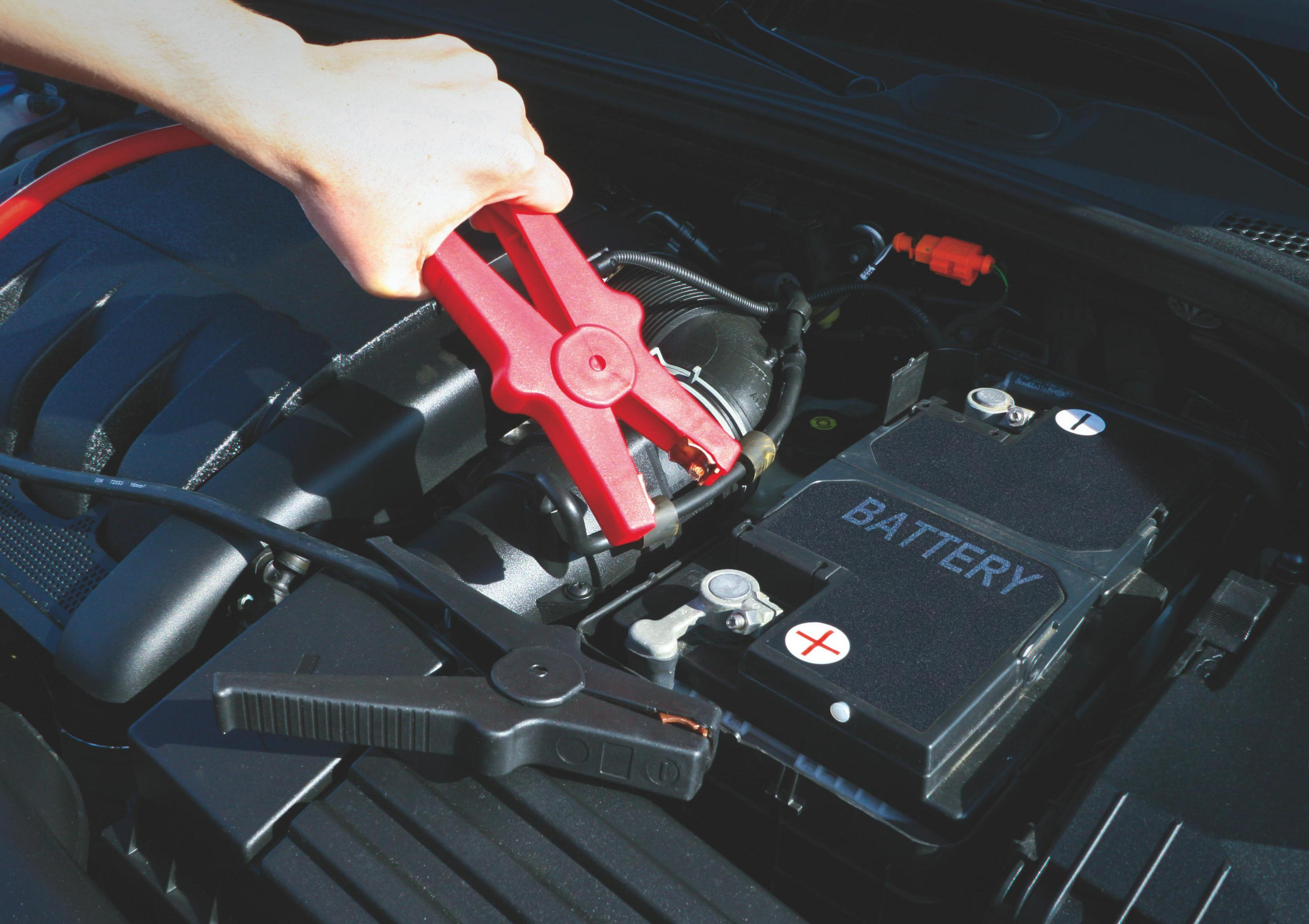Eine Hand verbindet rote Überbrückungskabel mit einer Autobatterie. Der Motor ist im Hintergrund.