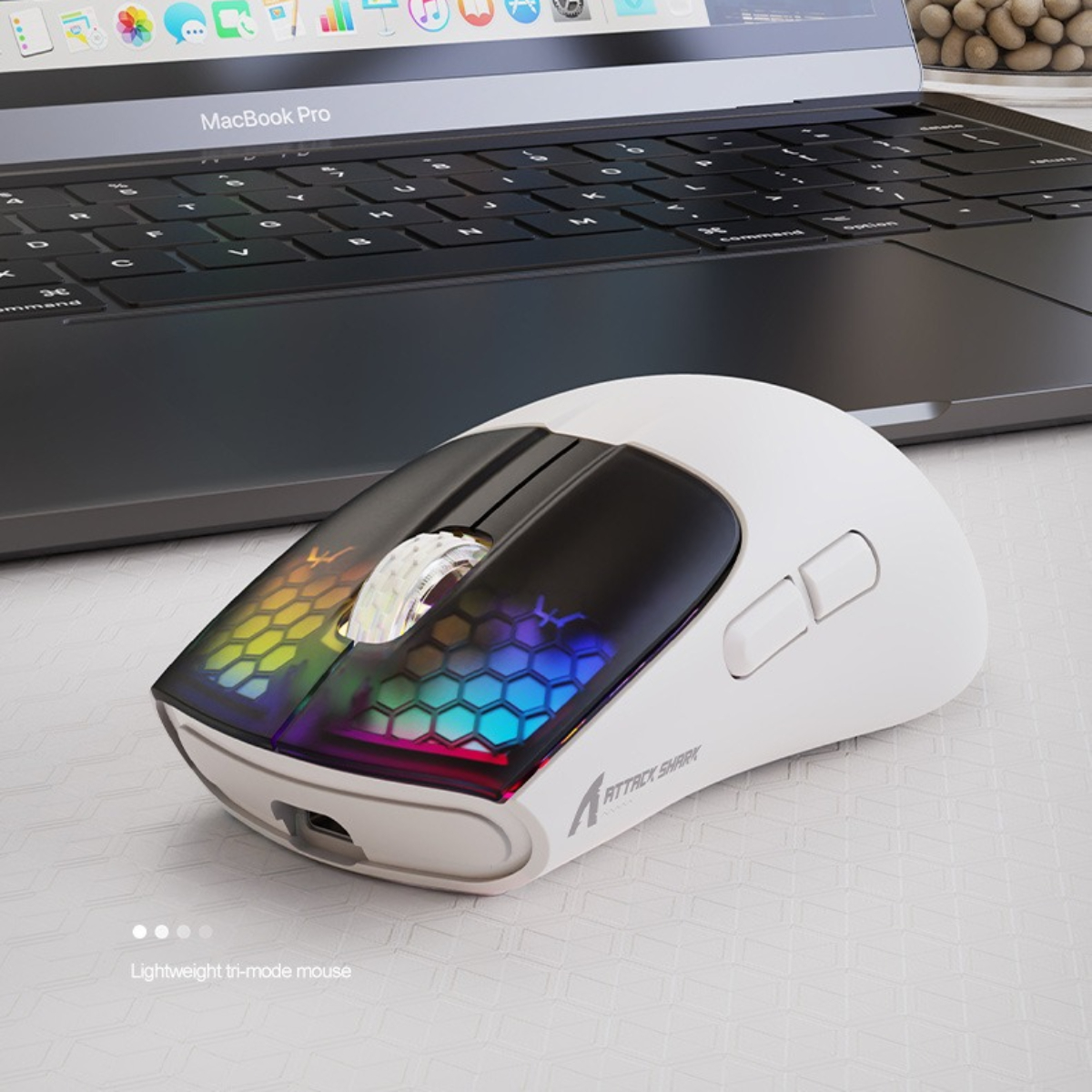 Eine weiße Gaming-Maus mit RGB-Beleuchtung. Sie befindet sich neben einem Laptop auf einer weißen Oberfläche.