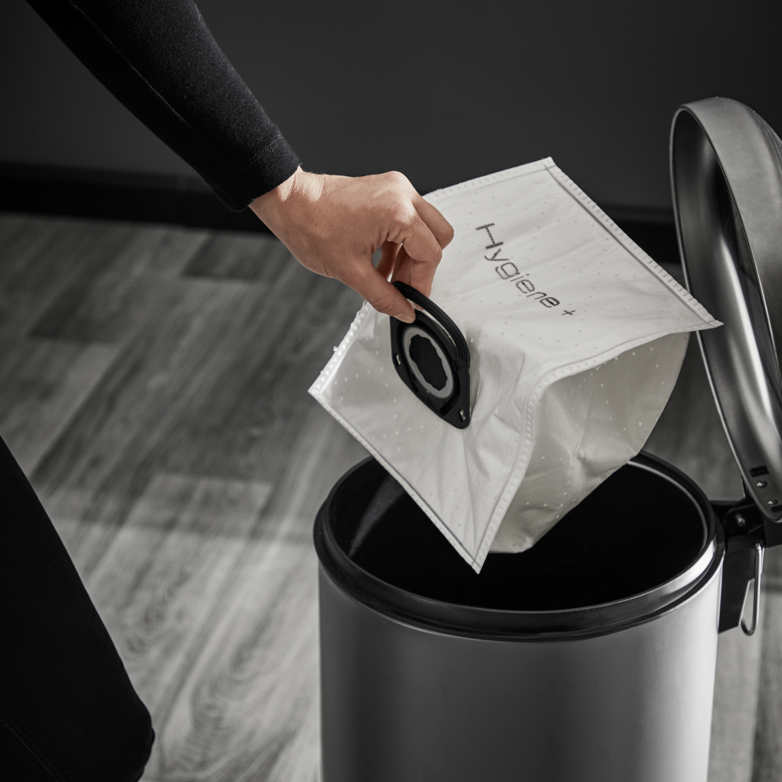 Ręka trzyma biały worek na śmieci nad szarym koszem na śmieci. Podłoga jest szara.