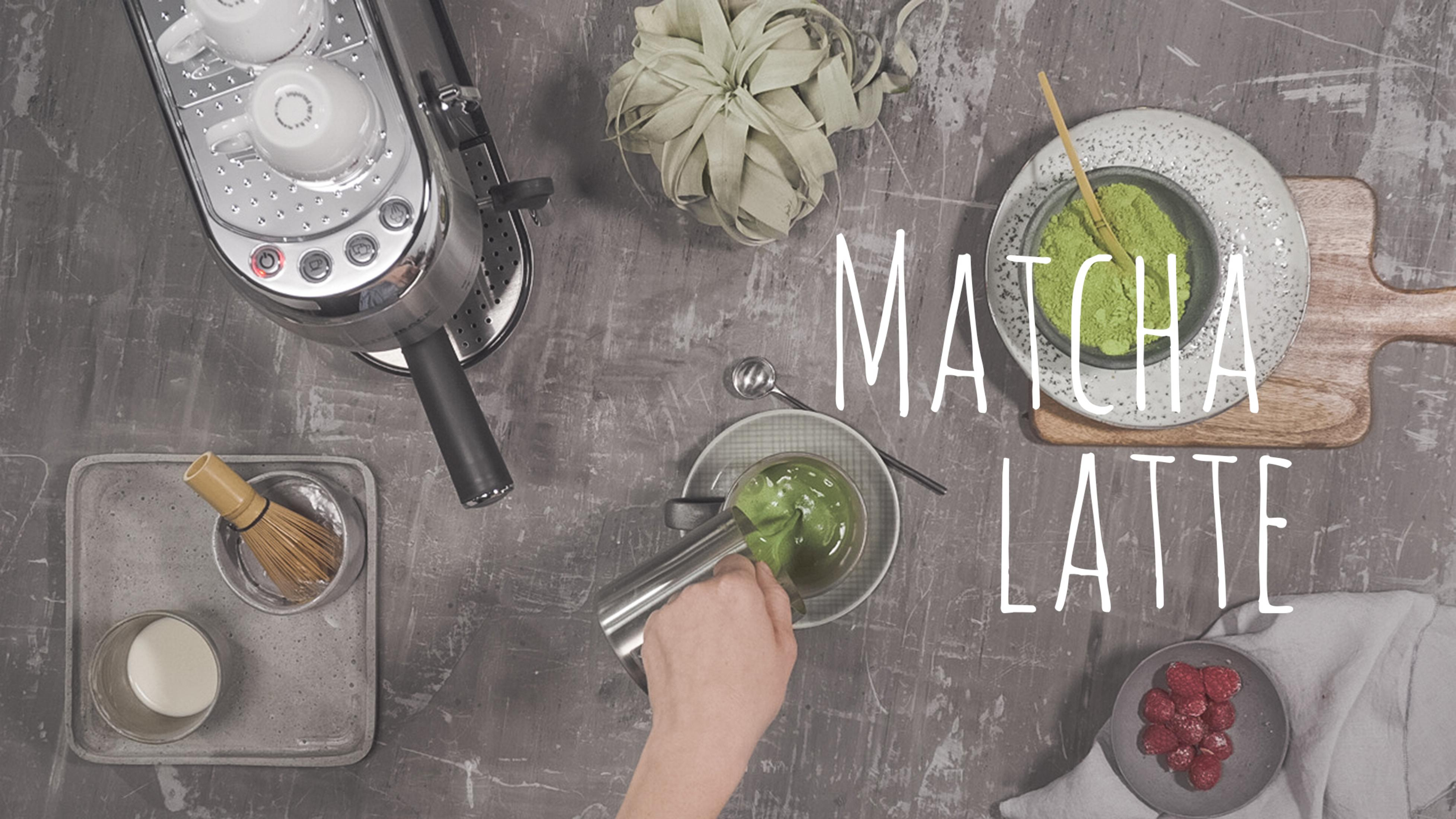 Matcha latte wlewana do filiżanki. Składniki i sprzęt widoczne na szarej powierzchni.