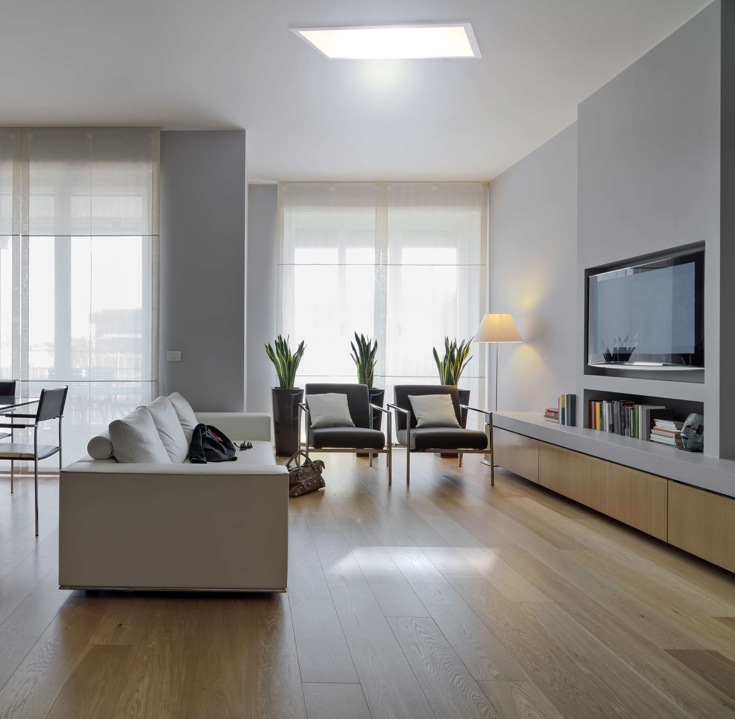Modernes Wohnzimmer mit Holzboden, weißem Sofa und großen Fenstern. Ein Fernseher an der Wand.