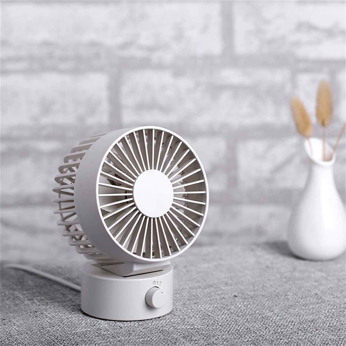 Ein weißer Ventilator auf grauer Oberfläche mit Vase und Zierpflanzen im Hintergrund.