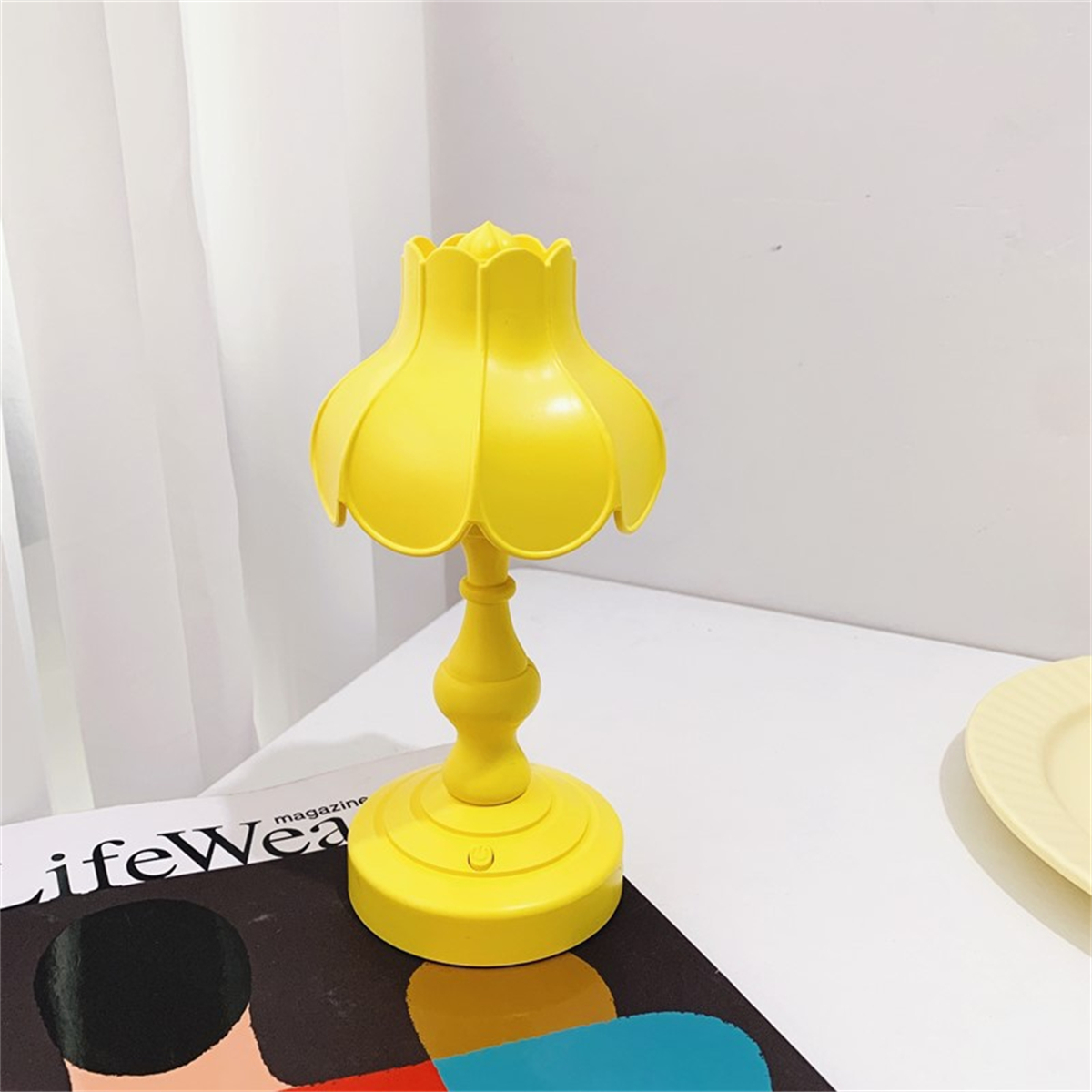 Eine gelbe Lampe mit blütenförmigem Schirm auf einer weißen Oberfläche. Sie hat einen dekorativen Fuß.