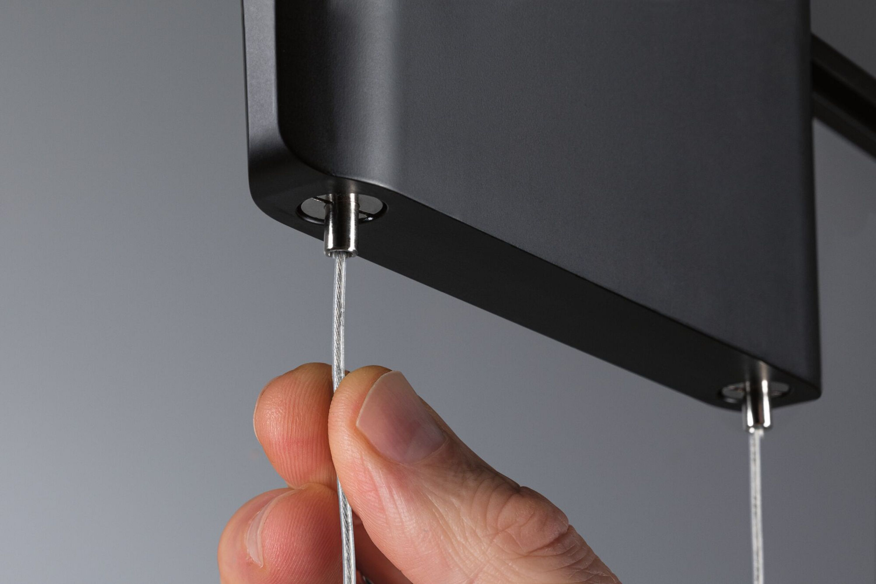 Hand hält ein dünnes, silbernes Kabel, das mit einem schwarzen Gerät verbunden ist, grauer Hintergrund.