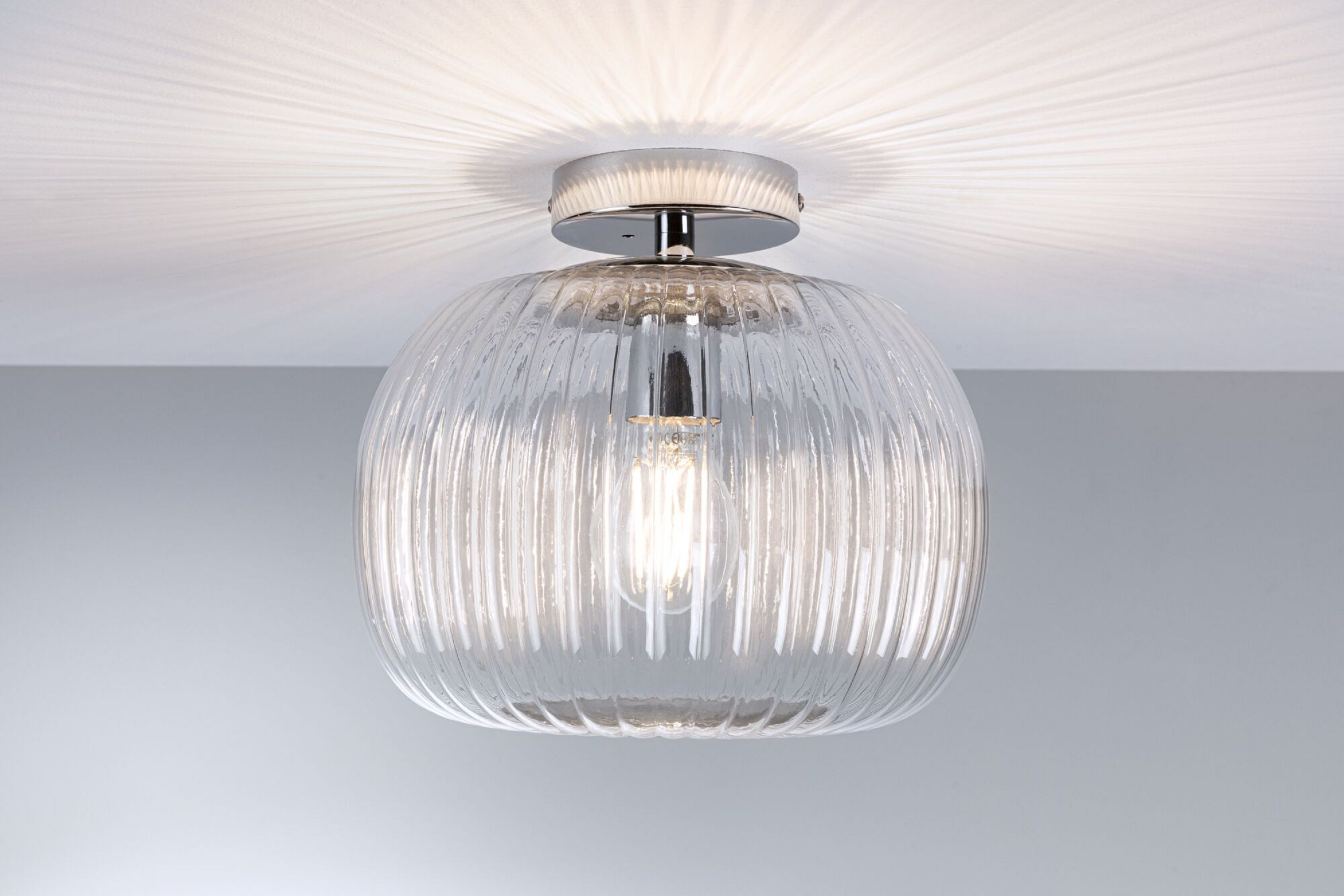 Lampa sufitowa z przezroczystym, żebrowanym szklanym kloszem i chromowaną podstawą. Światło jest włączone.