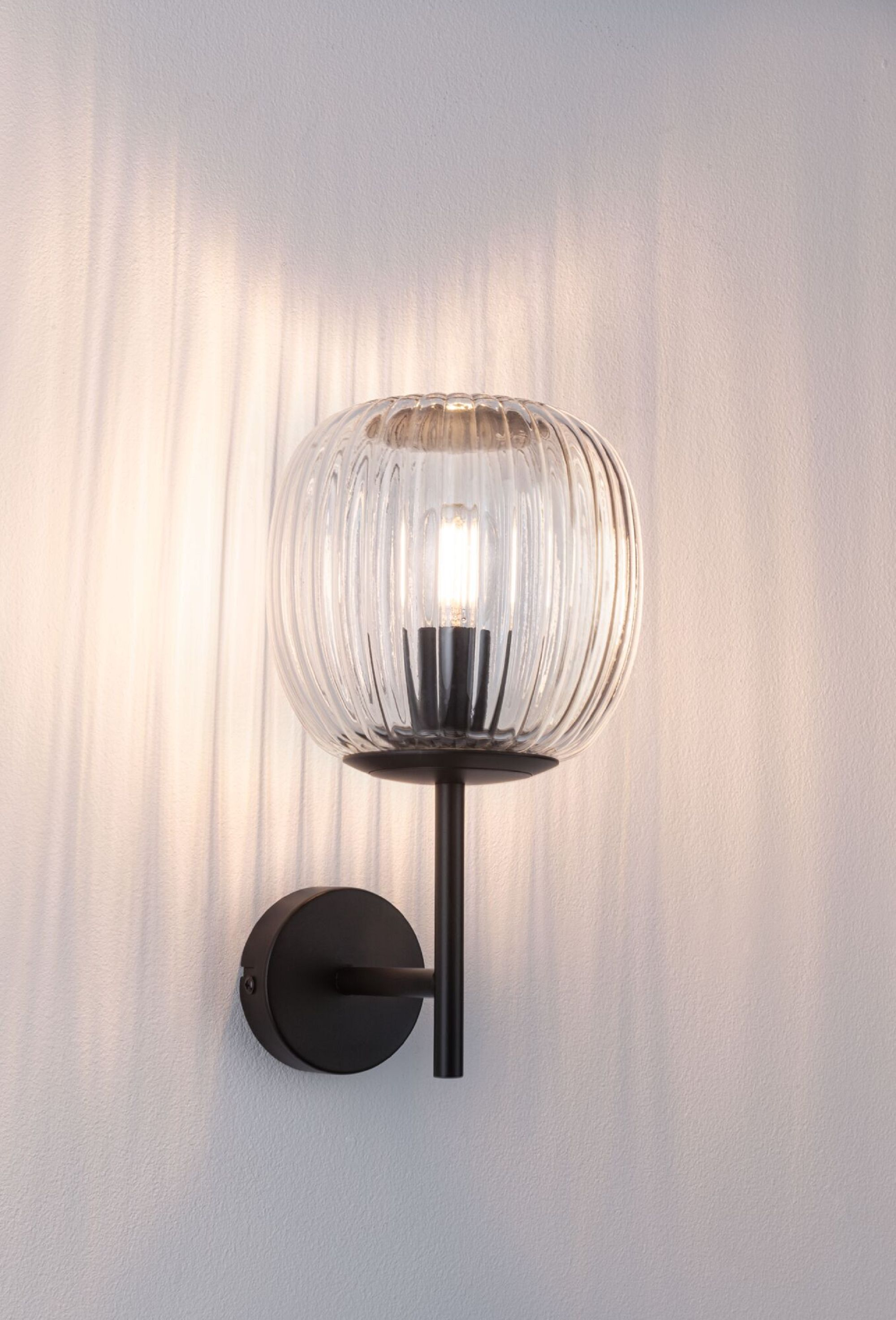 Eine schwarze Wandlampe mit einem klaren, gerippten Glasschirm, der Schatten wirft.