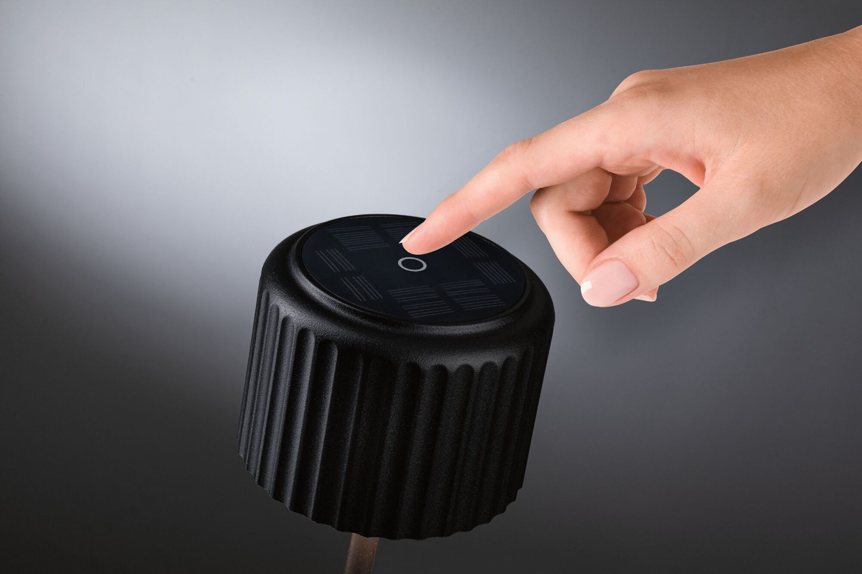 Dłoń naciska przycisk na czarnej, cylindrycznej lampie solarnej ogrodowej.