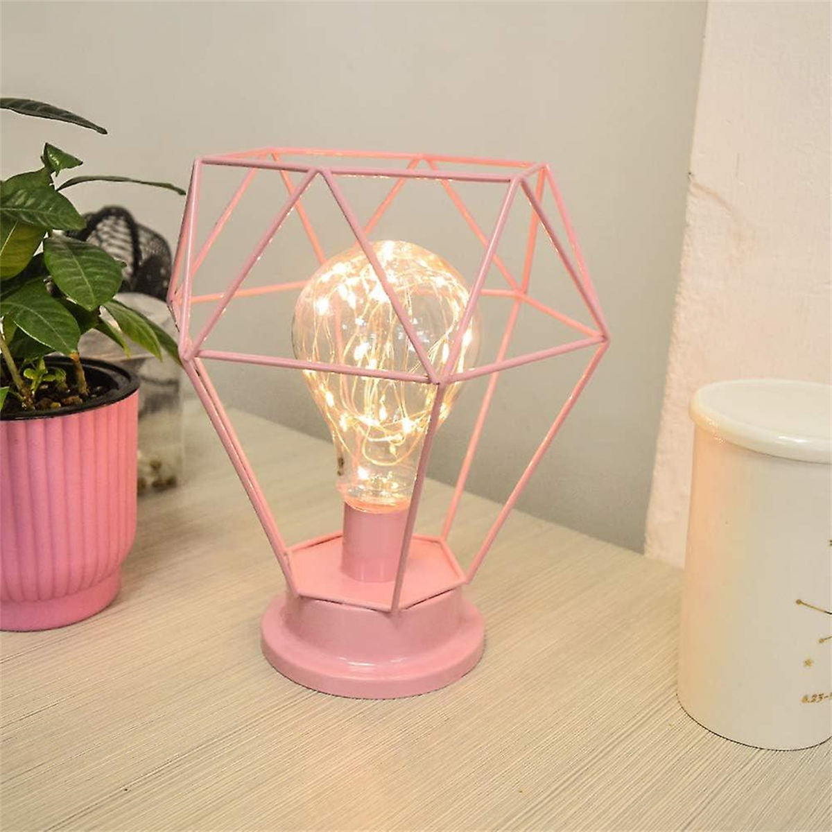 Rosa geometrische Käfiglampe mit Glühbirne. Rosa Hintergrund. Eine weiße Tasse ist ebenfalls vorhanden.