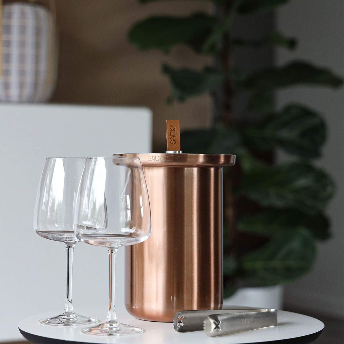 Weingläser und Kupfer-Weinkühler auf einem weißen Tisch. Im Hintergrund ist eine Pflanze sichtbar.