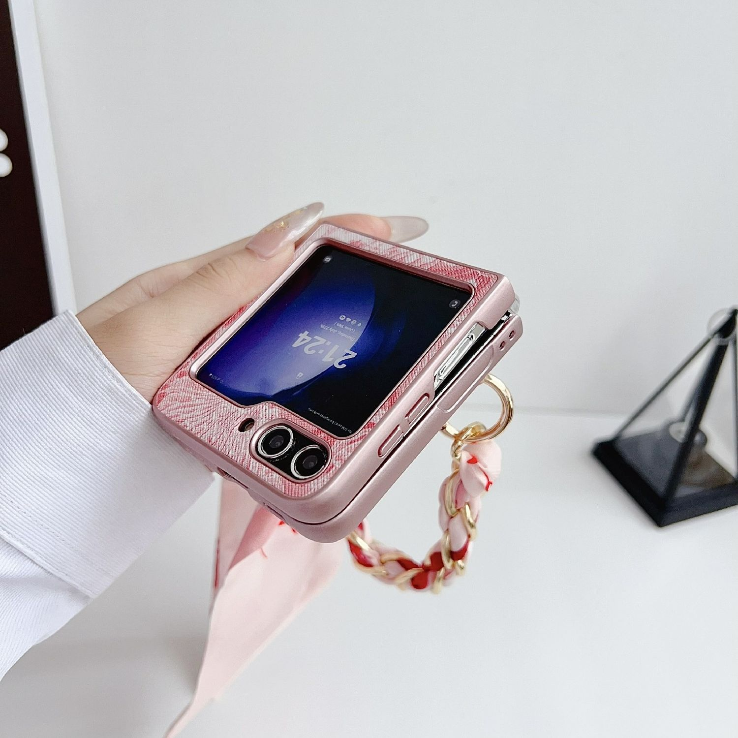 Mano sosteniendo una funda rosa. La funda tiene una cadena. El teléfono está abierto. Fondo blanco.