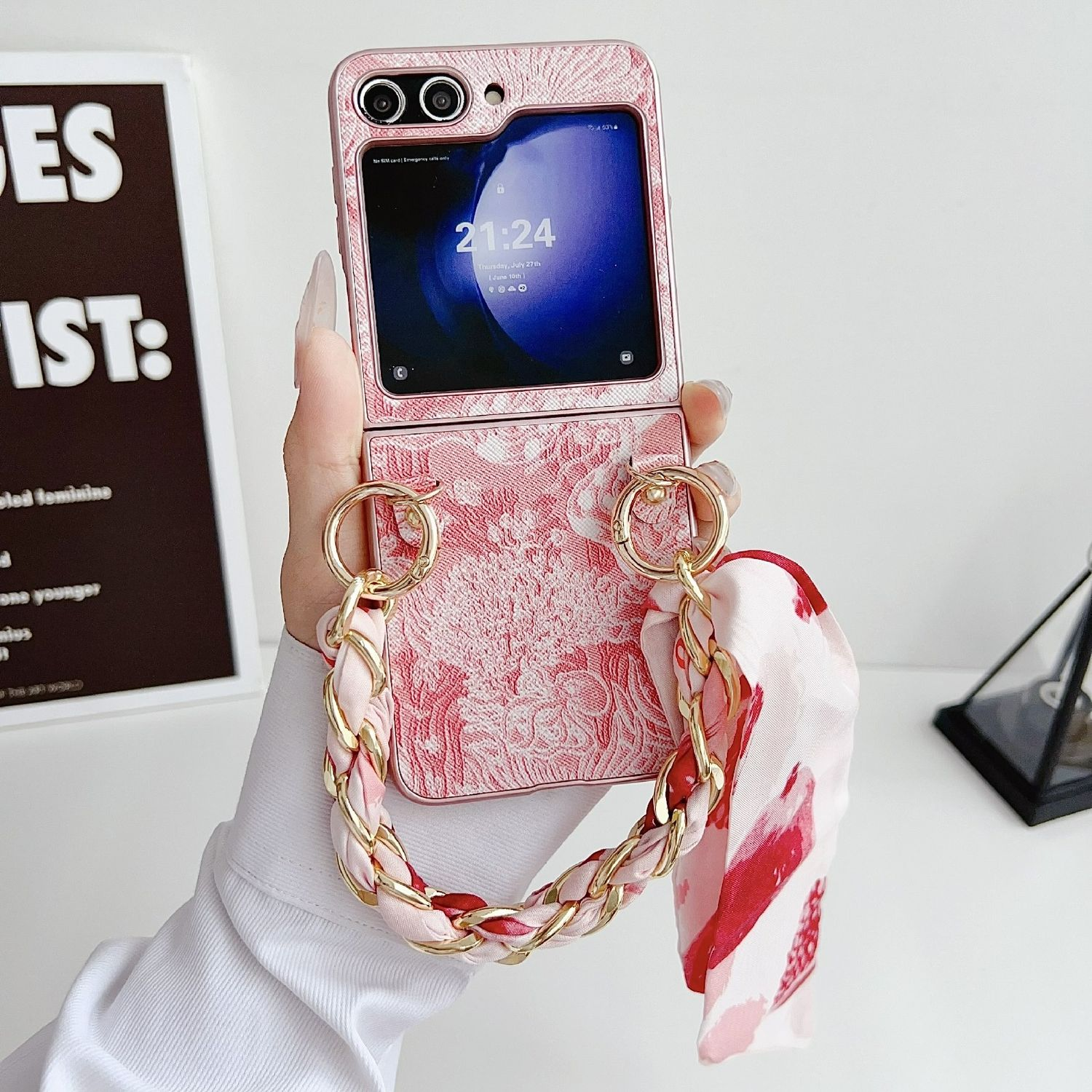 Funda de teléfono rosa con correa de cadena sostiene un teléfono plegado que muestra la hora. Fondo: pared blanca.