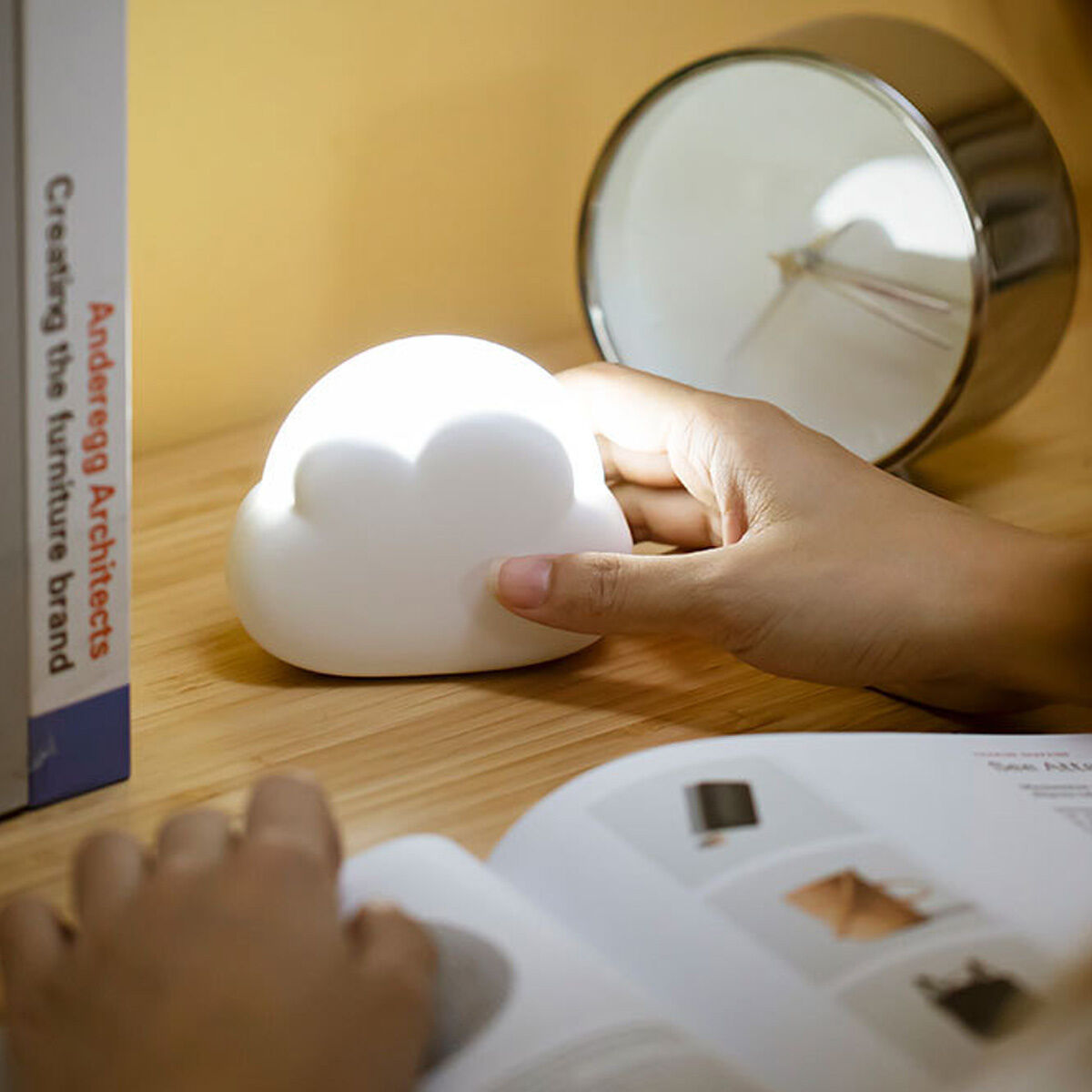 Eine wolkenförmige Lampe leuchtet auf einem Holztisch, neben einem Buch und einer Uhr.