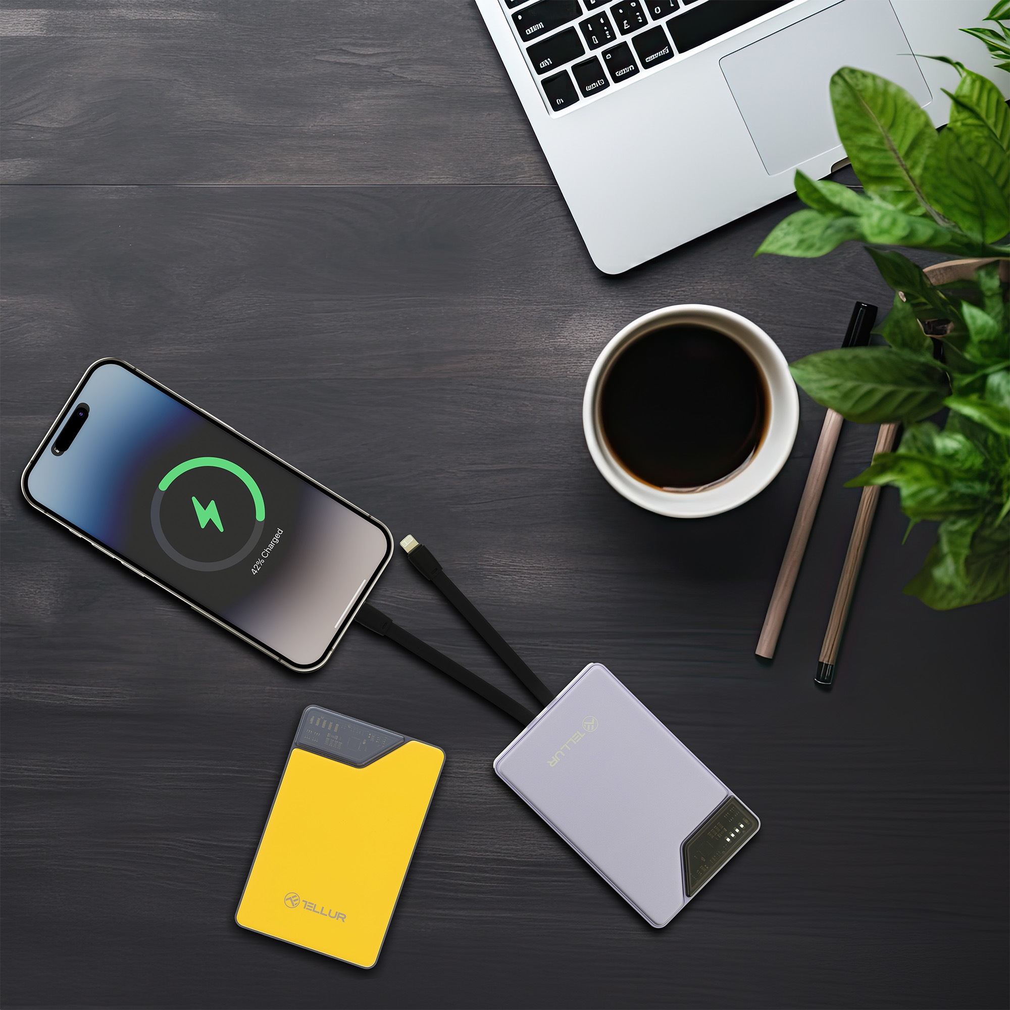 Telefon ładujący się bezprzewodowo, obok laptopa, kawy i dwóch powerbanków na ciemnym drewnianym stole.