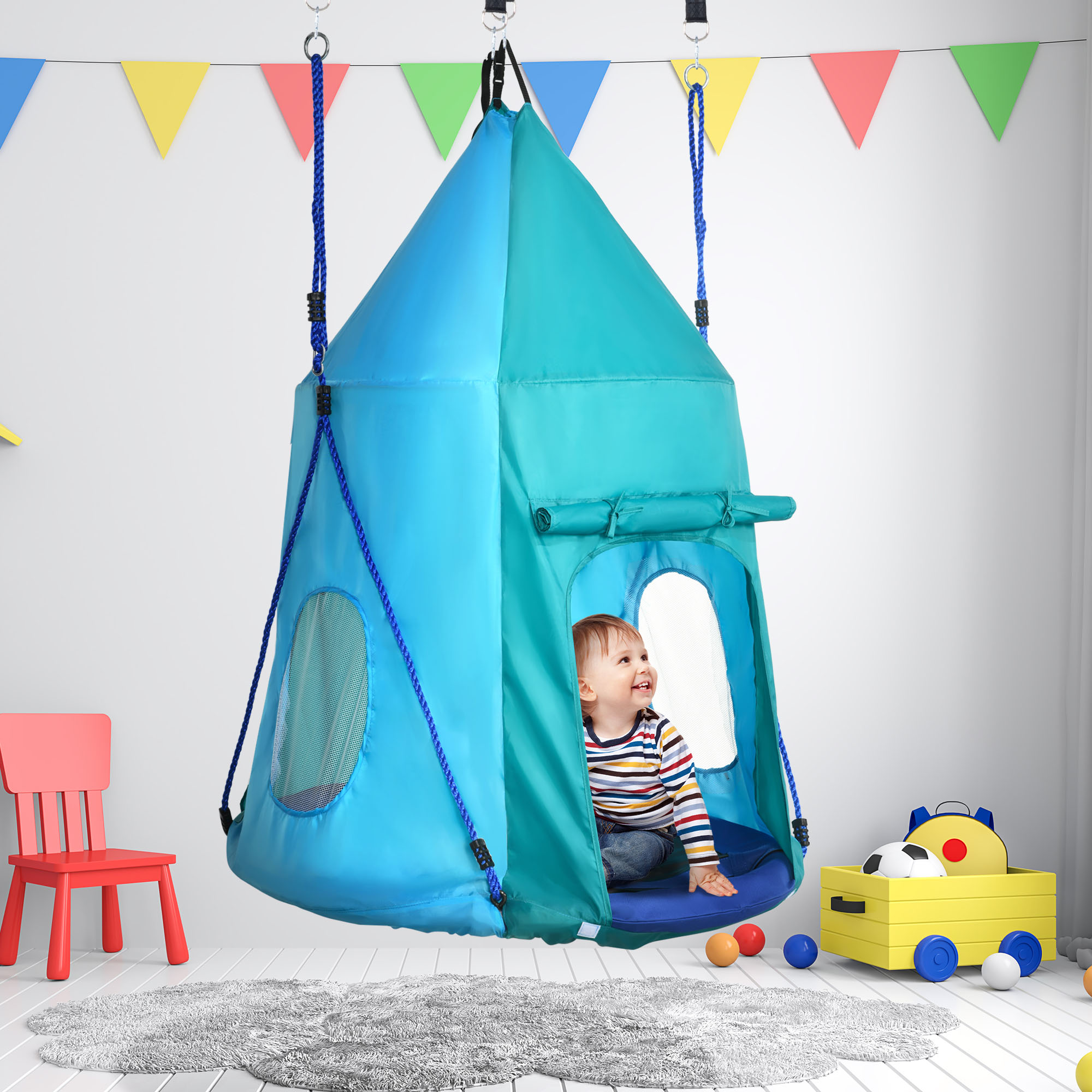Bambino in un'altalena-tenda blu e verde. Striscioni colorati sullo sfondo. Stanza bianca.