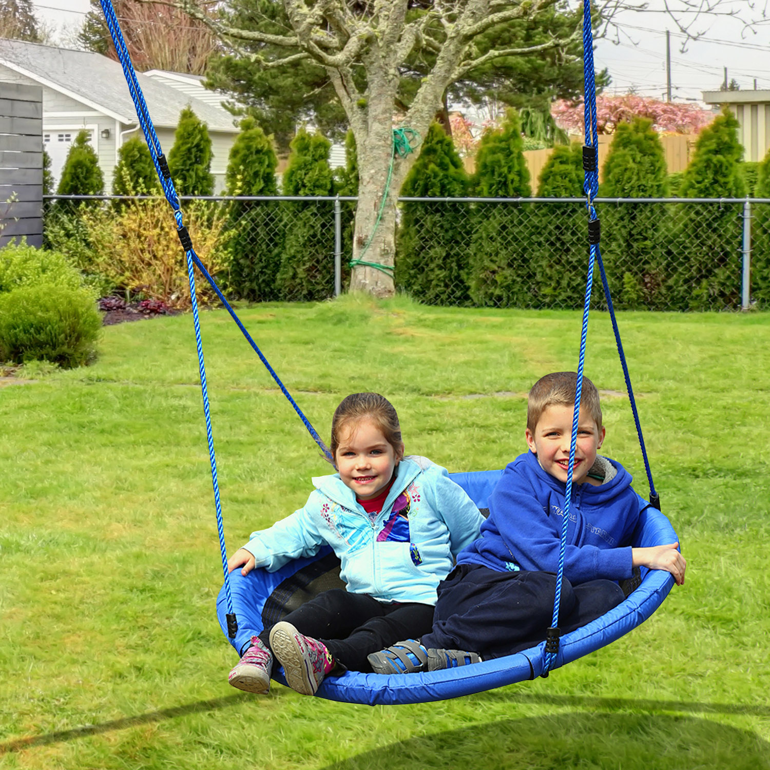 Due bambini su un'altalena blu, erba verde e alberi sullo sfondo.