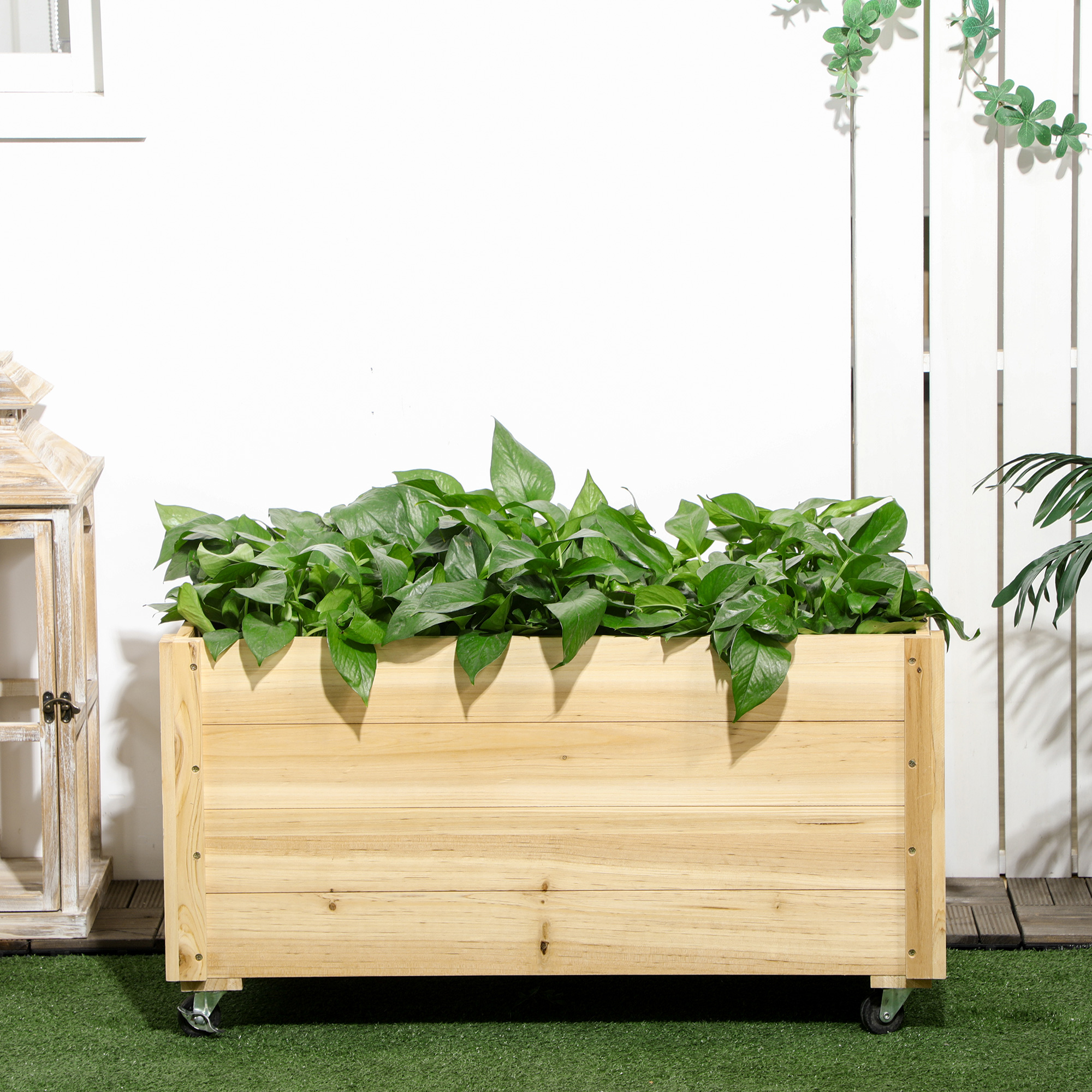 Fioriera in legno su ruote, piena di piante verdi, su superficie verde.