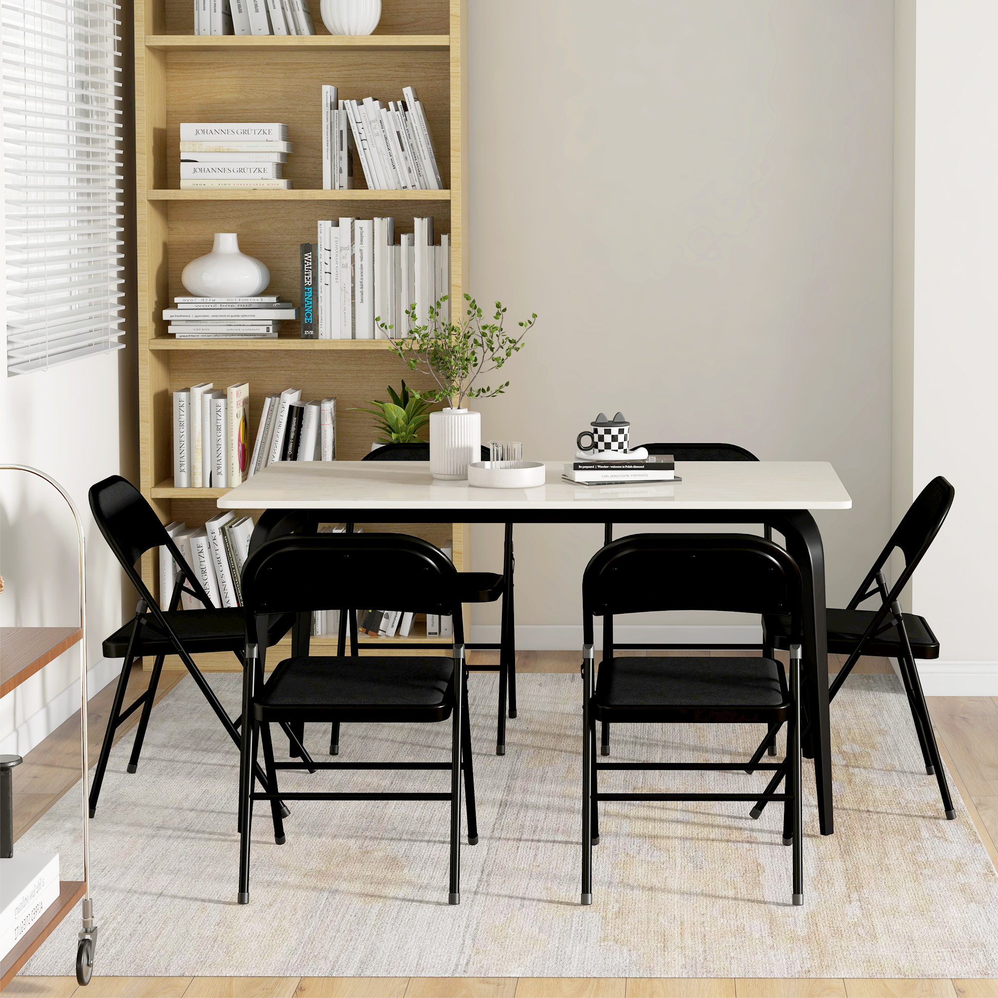 Tavolo da pranzo con quattro sedie nere. Libreria, parete bianca, finestra con tende.