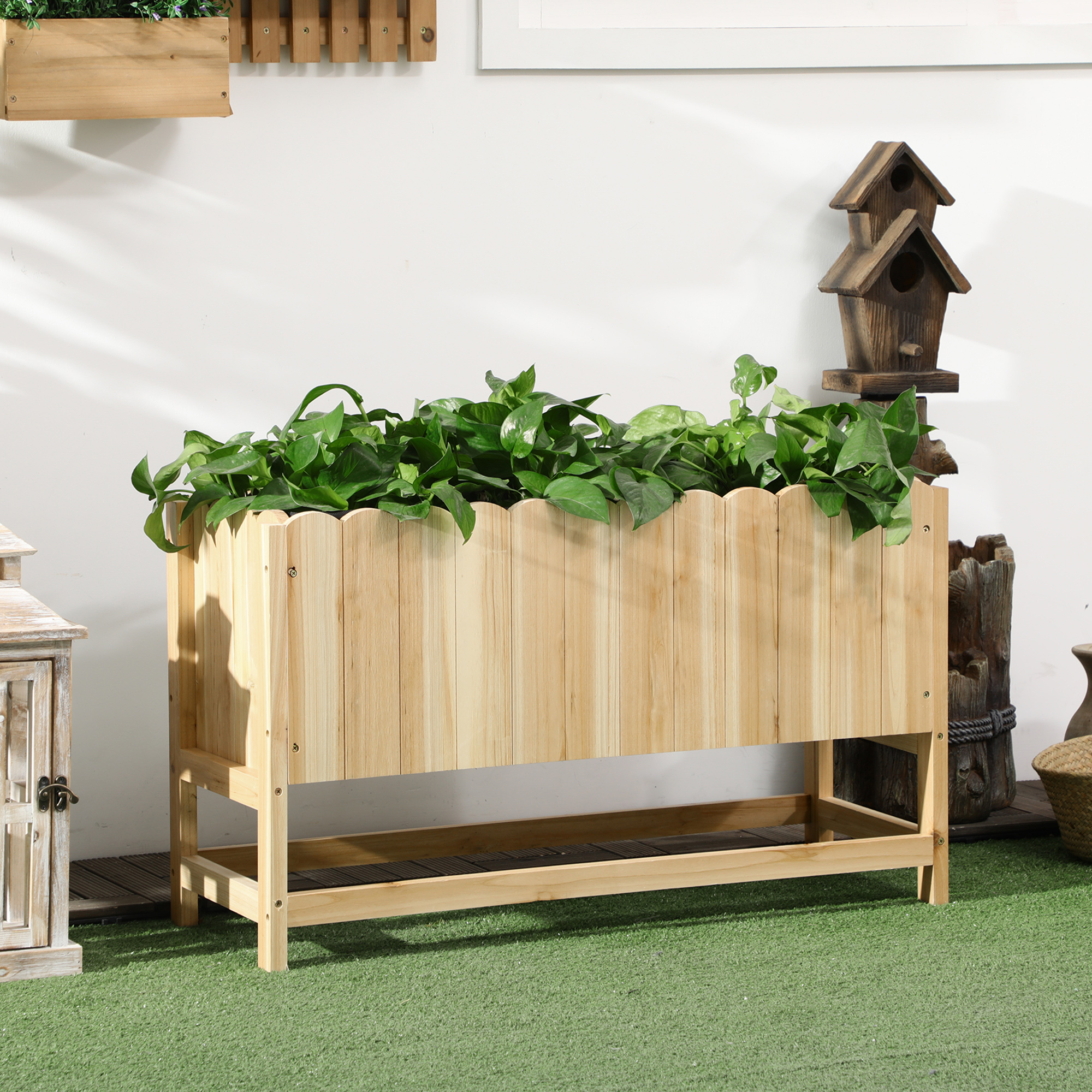 Fioriera in legno con piante, su erba verde, con una casetta per uccelli e un muro bianco sullo sfondo.