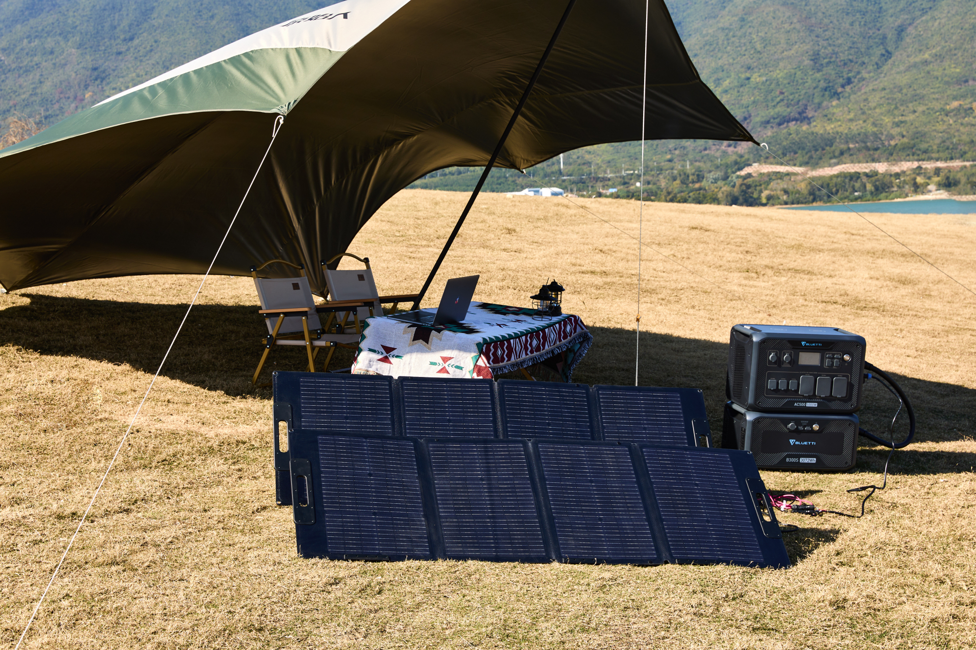Una tenda con tavolo, laptop e pannelli solari è allestita all'aperto vicino a un lago e colline.