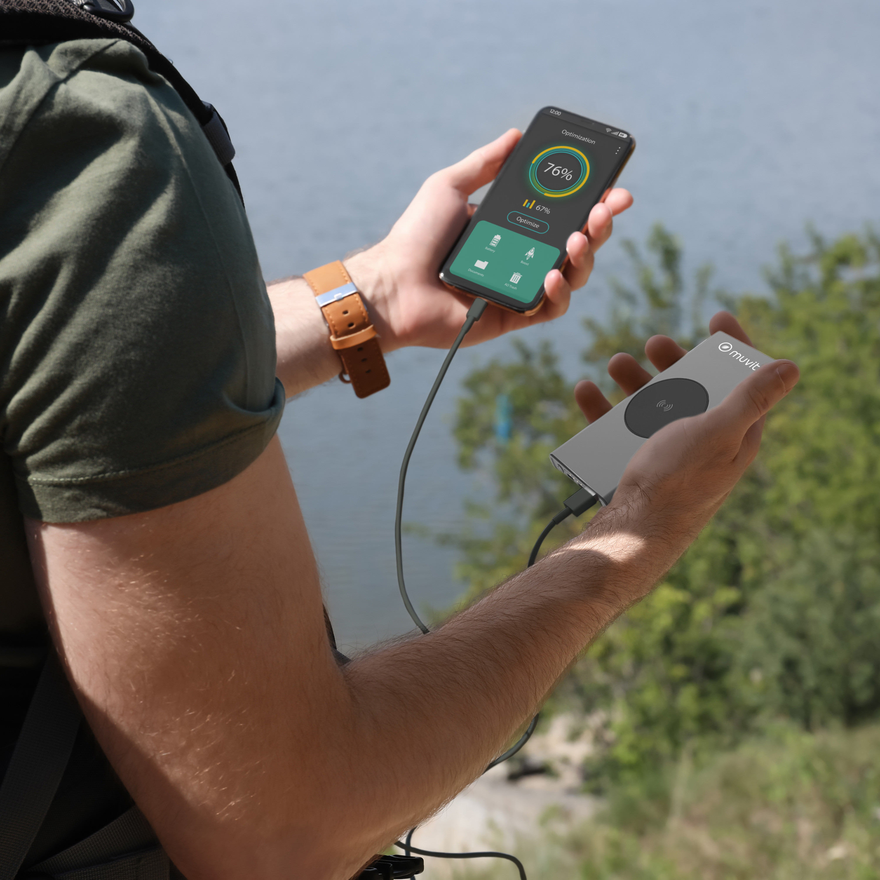 Persona sosteniendo un smartphone y un cargador inalámbrico. El teléfono muestra el porcentaje de batería. El fondo es un lago.