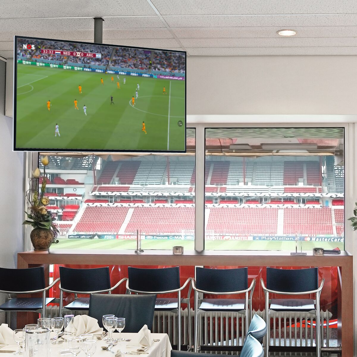 A TV shows a soccer match. People sit at tables, watching.  The background is a stadium.