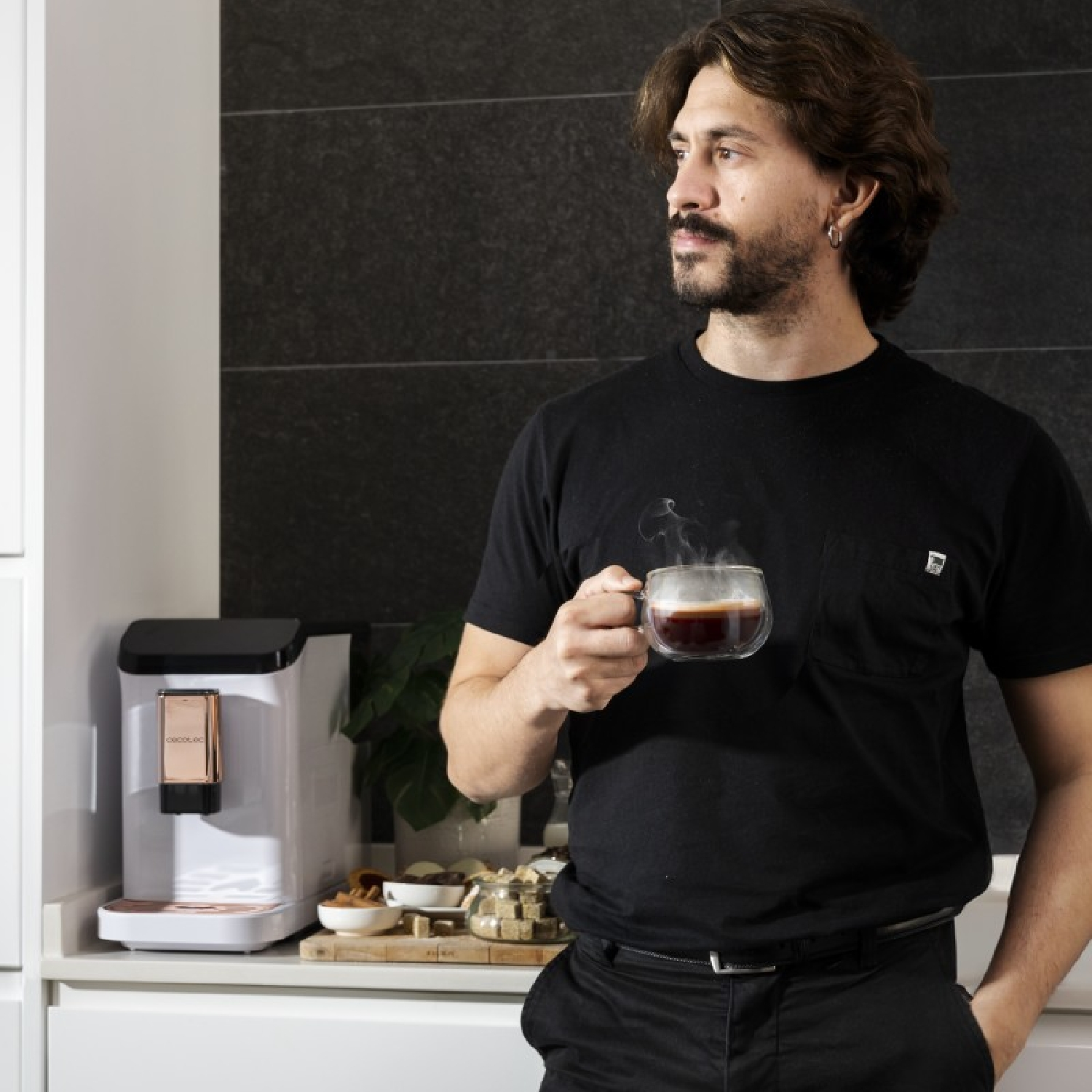 Hombre sosteniendo vaso de café, con cafetera y aperitivos cerca.