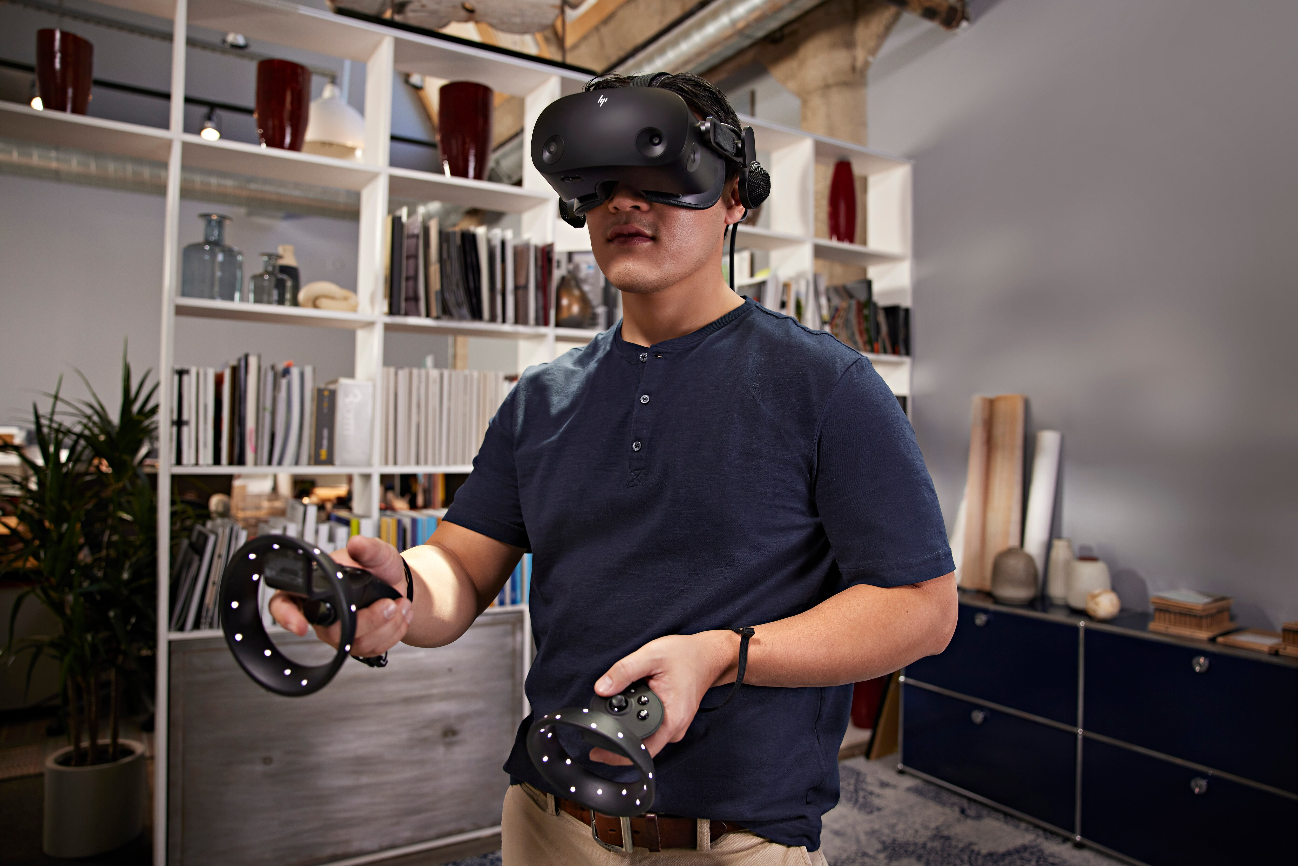 Mann mit VR-Headset und Controllern. Bücherregale und ein Schreibtisch im Hintergrund.