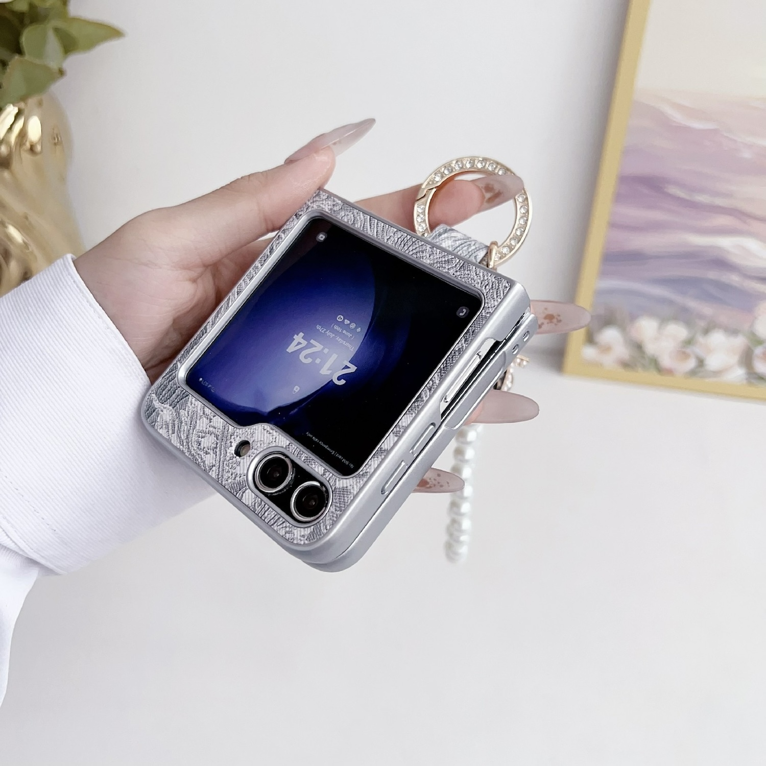 Hand, die ein silbernes faltbares Telefon in einer Schutzhülle hält, mit einem perlenbesetzten Anhänger.