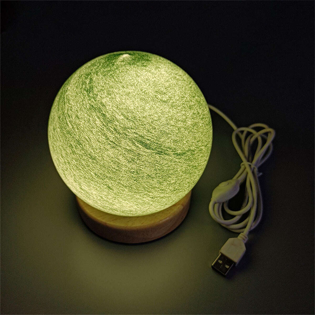 Mondförmige Lampe mit grünlicher, gesprenkelter Oberfläche, auf Holzsockel, mit USB-Kabel.