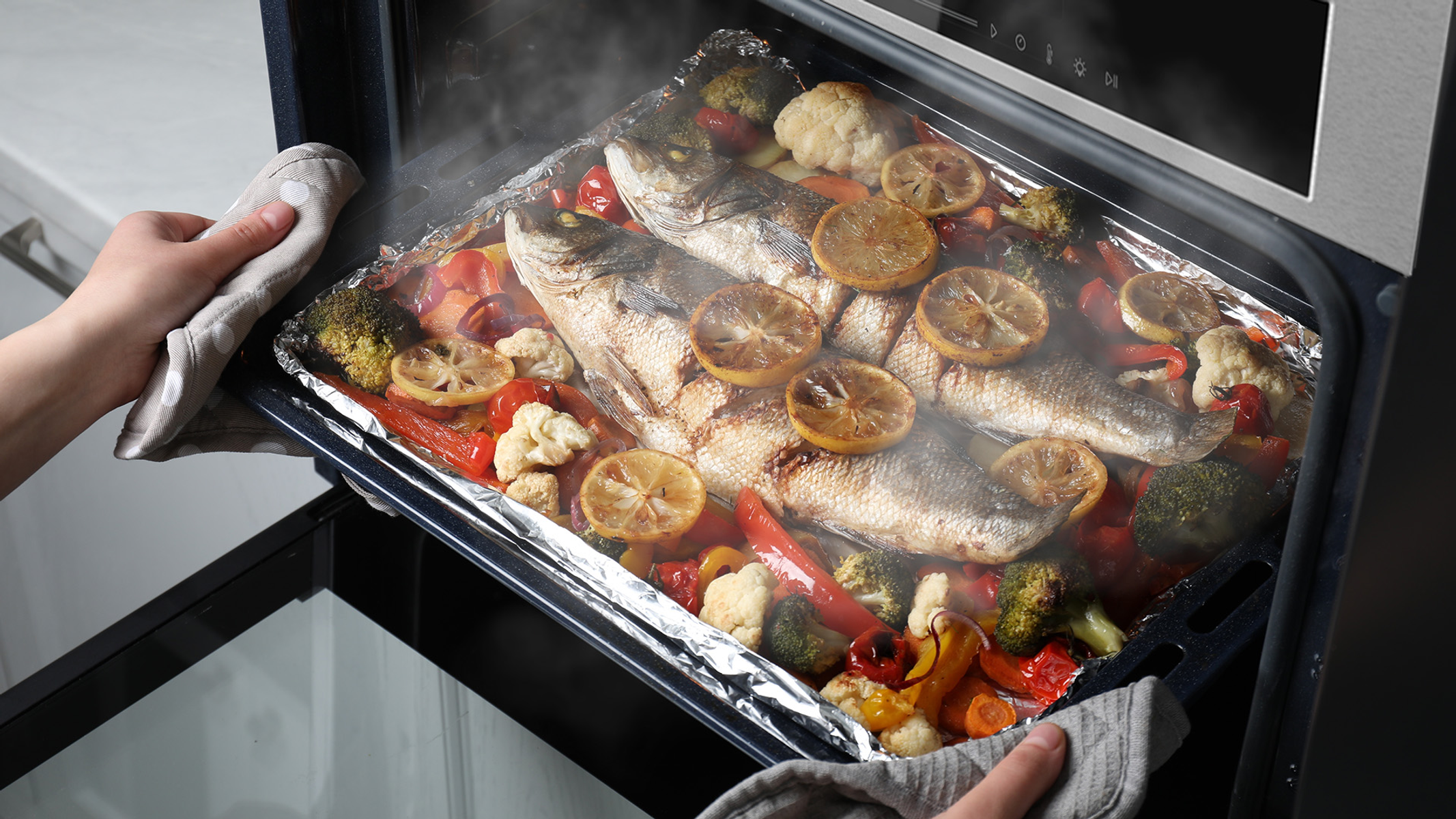 Horno con pescado y verduras cocinados. Manos con guantes de horno sostienen la bandeja.