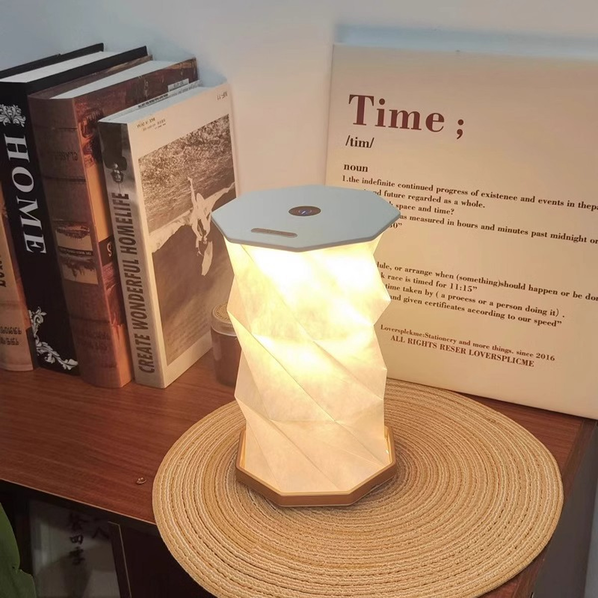 Eine moderne Lampe im Origami-Stil auf Holzsockel, neben Büchern und einem gerahmten Zitat.