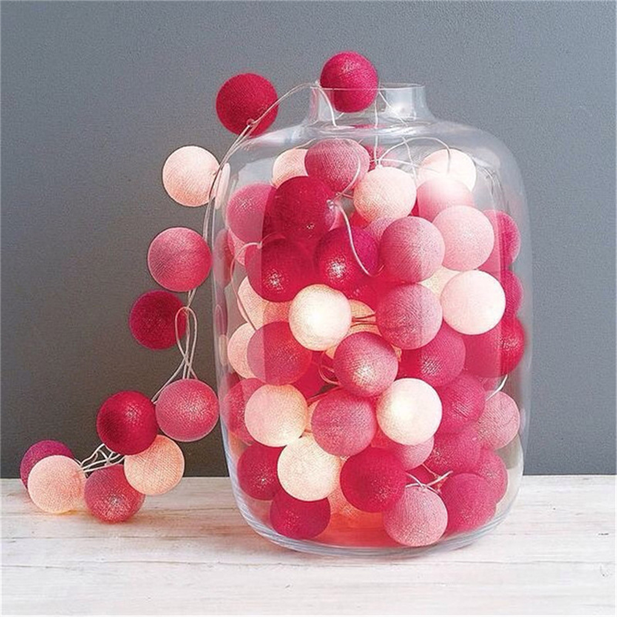 Glasbehälter gefüllt mit roten und rosa Baumwollkugel-Lichterketten, Holzoberfläche, graue Wand.