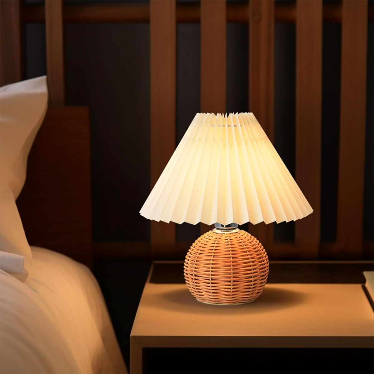 Eine beleuchtete Nachttischlampe mit plissiertem Lampenschirm auf einem Nachttisch neben einem Bett.