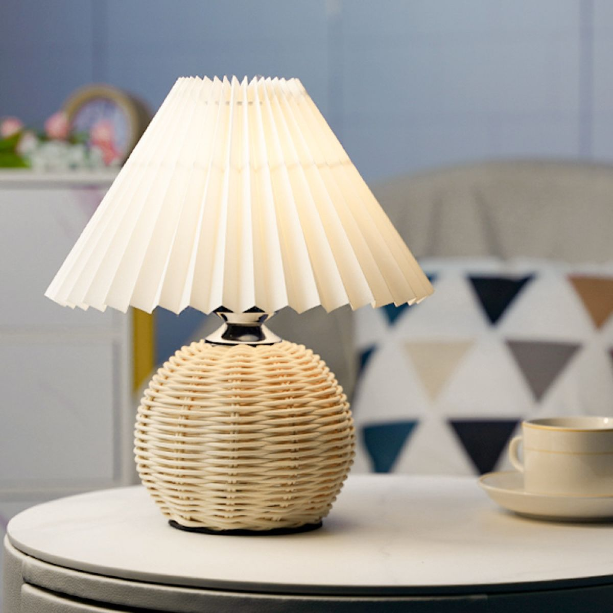 Eine Tischlampe mit plissiertem Lampenschirm und gewebtem Sockel auf einem Tisch. Rechts steht eine Tasse.