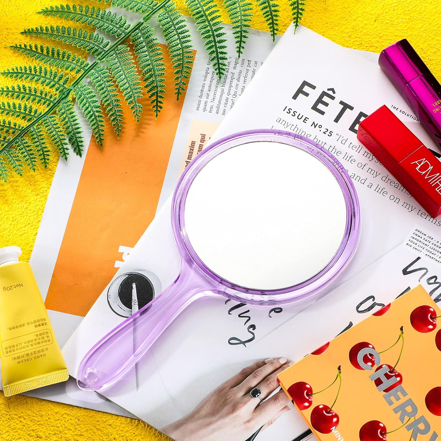 Eine Beauty-Szene auf Gelb. Handspiegel, Lippenstift, Creme, Zeitschrift und eine Hand mit Ring sind sichtbar.