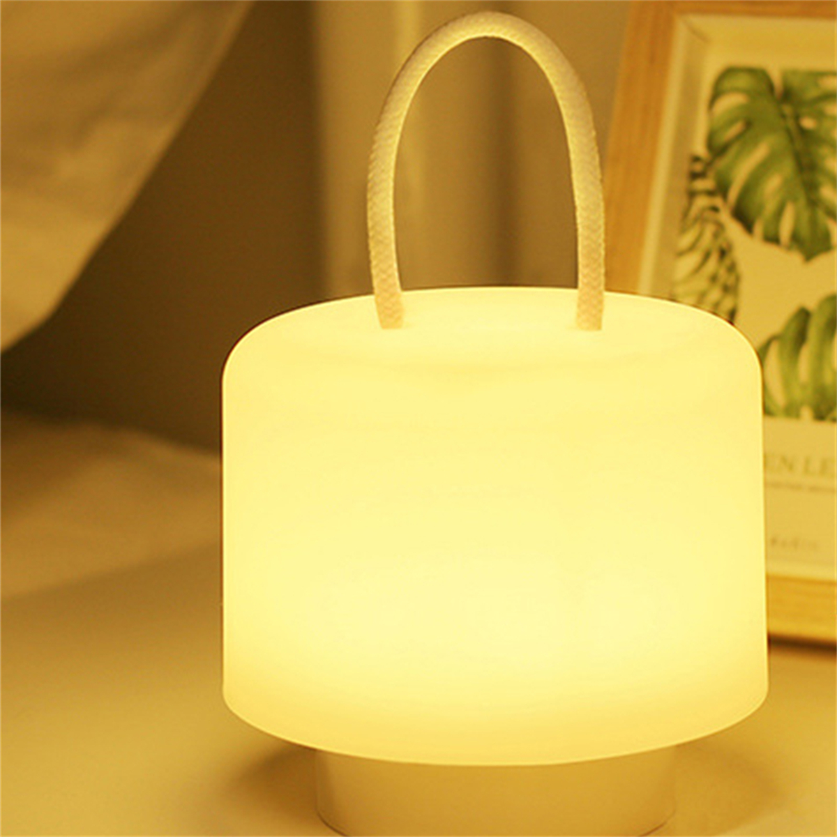Eine leuchtende weiße Nachttischlampe mit Griff, auf einem Tisch. Im Hintergrund ein Bilderrahmen.