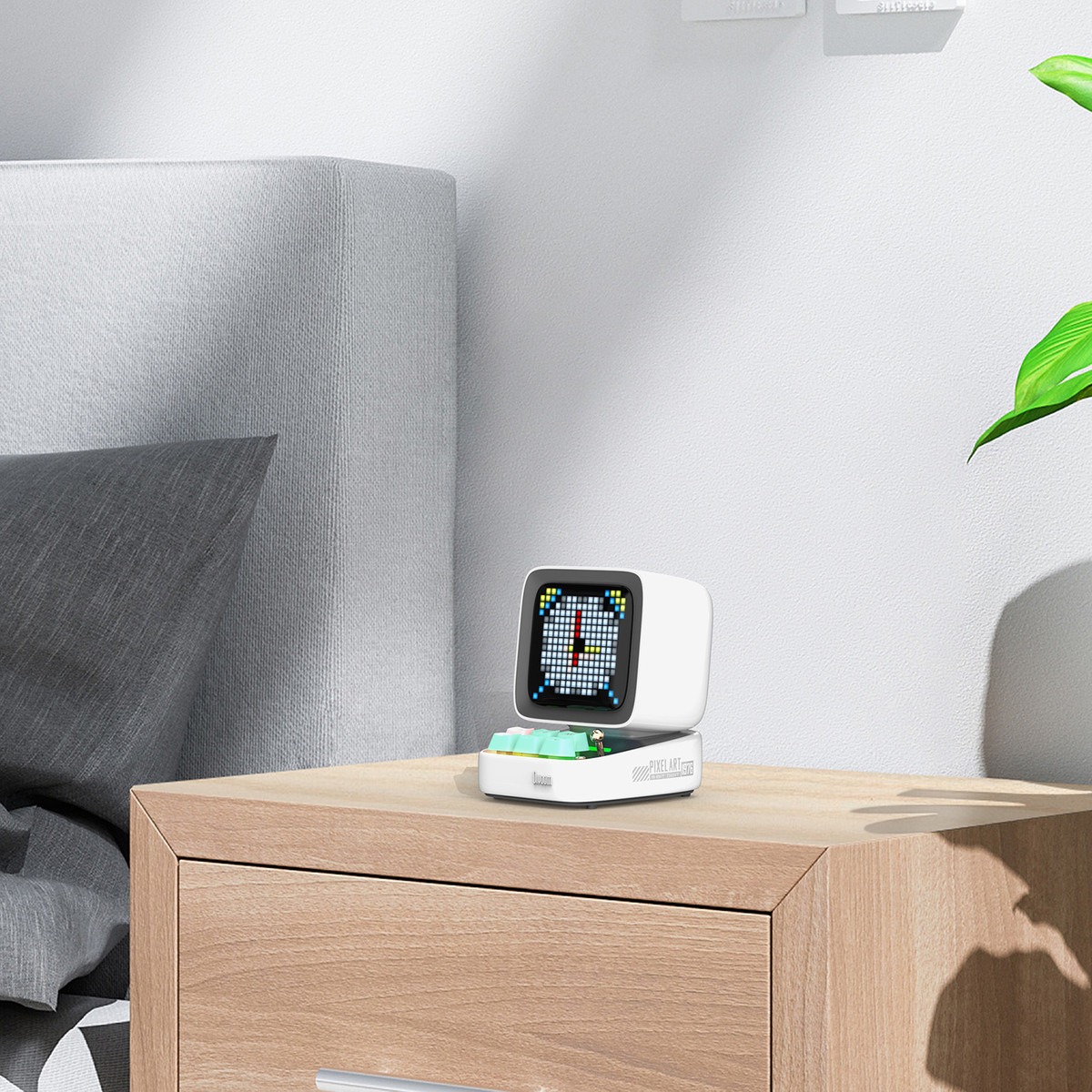 Biały głośnik z wyświetlaczem pixel art, na drewnianej szafce nocnej, z zieloną rośliną.