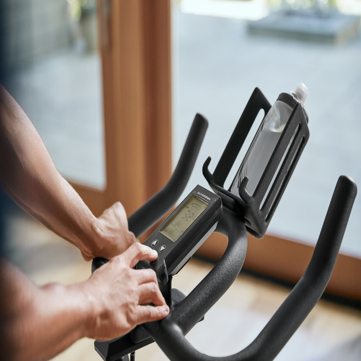 Ręce na konsoli roweru stacjonarnego. Zamontowana butelka wody. Tło to okno.