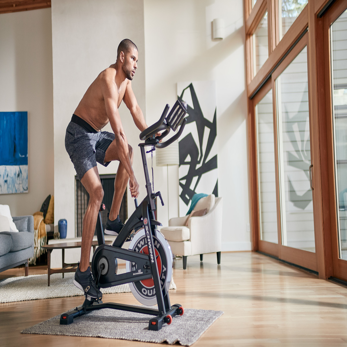 Mężczyzna na rowerze stacjonarnym. W pomieszczeniu, bez koszuli. Nowoczesny salon z dużym oknem. Skupiony.
