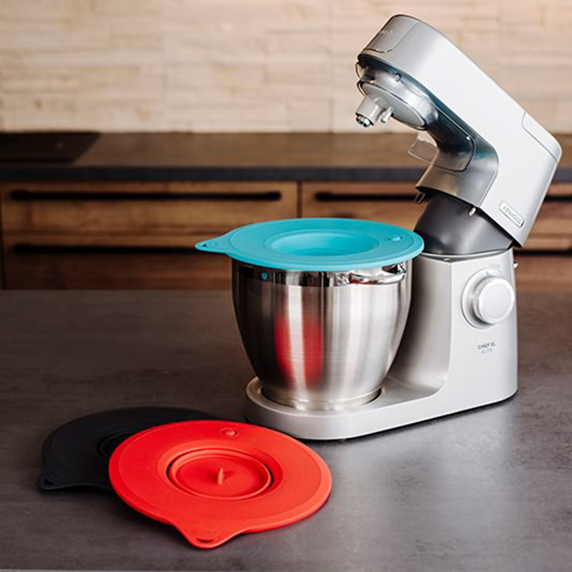 Ein silberner Küchenmixer mit blauem Deckel und zwei farbigen Deckeln auf einem grauen Tisch.