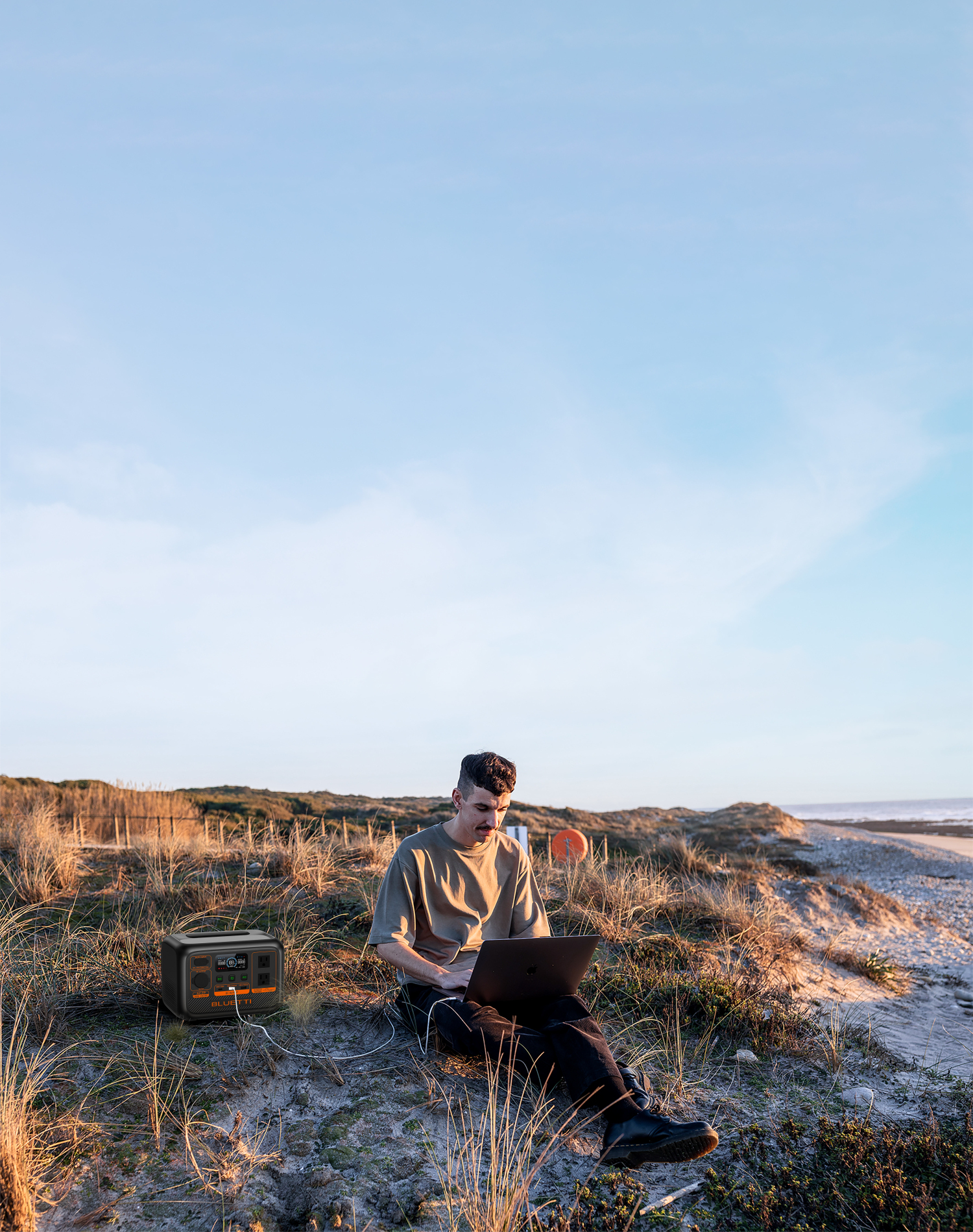 Mężczyzna siedzi na ziemi, pracując na laptopie. Stacja zasilania obok niego, na plaży z błękitnym niebem.