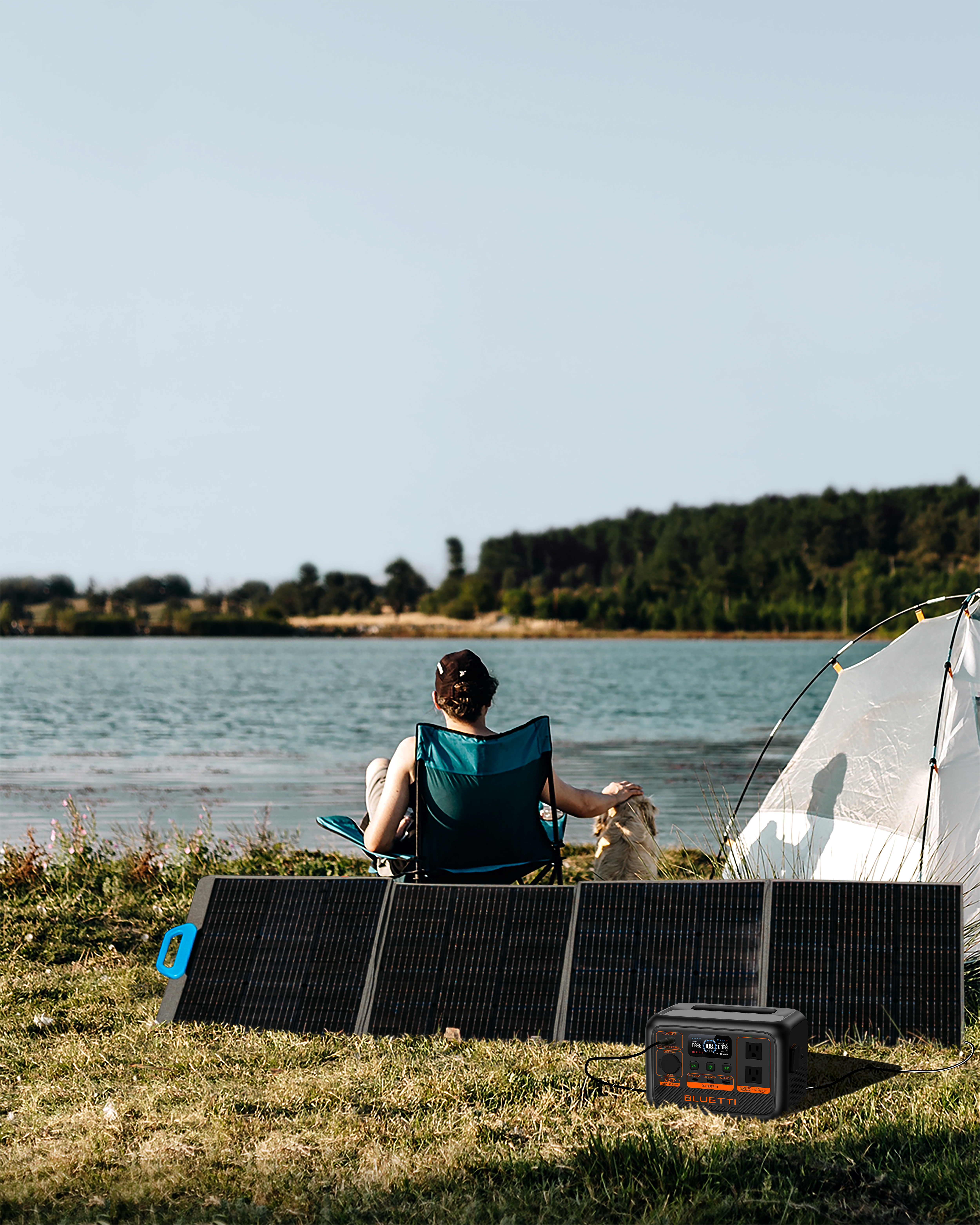 Osoba siedzi nad jeziorem, panel słoneczny i elektrownia widoczne. Namiot rozbity. Pies.
