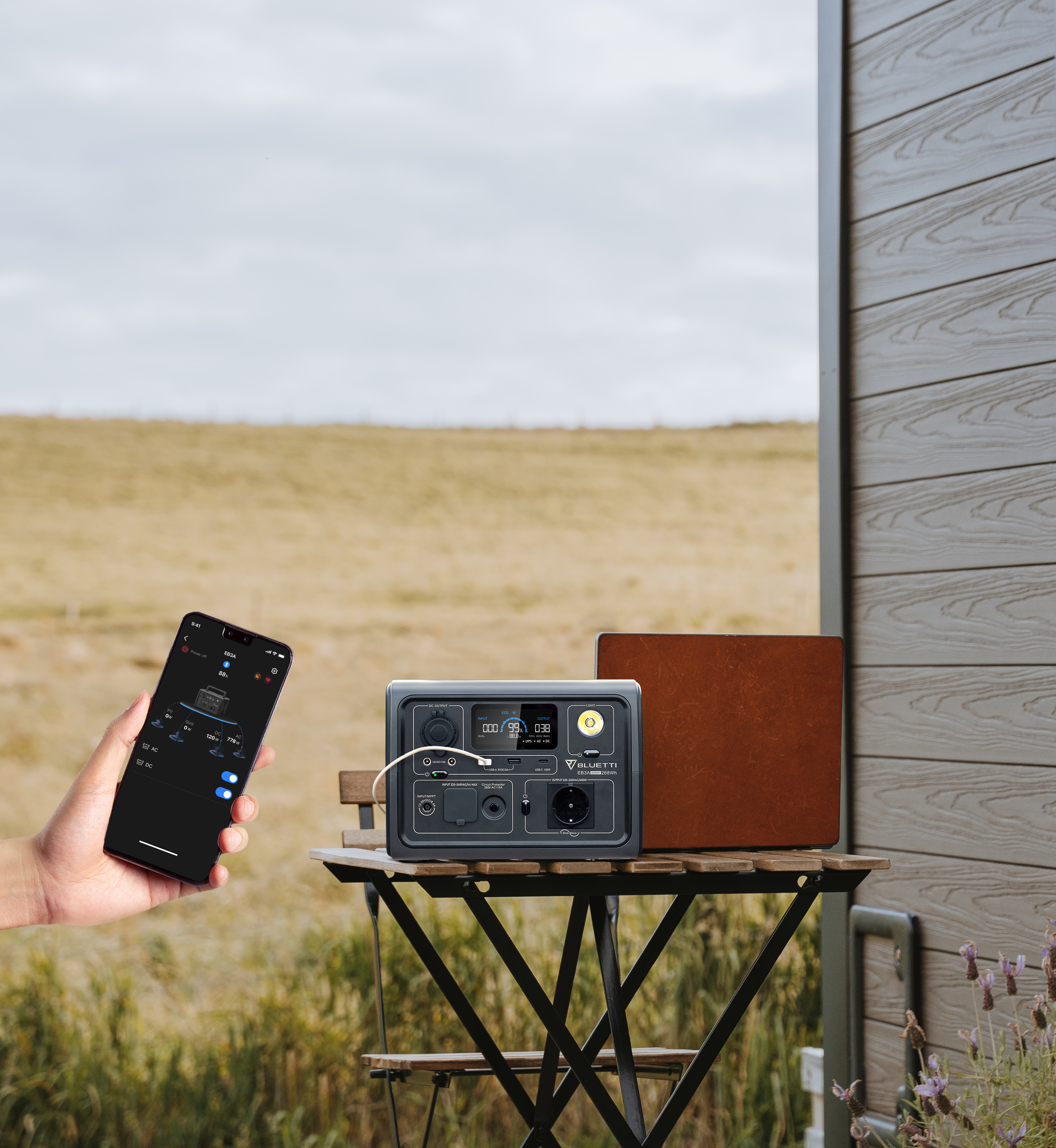 Person hält Telefon mit App, tragbare Powerstation und braune Box auf einem Tisch. Szene im Freien.