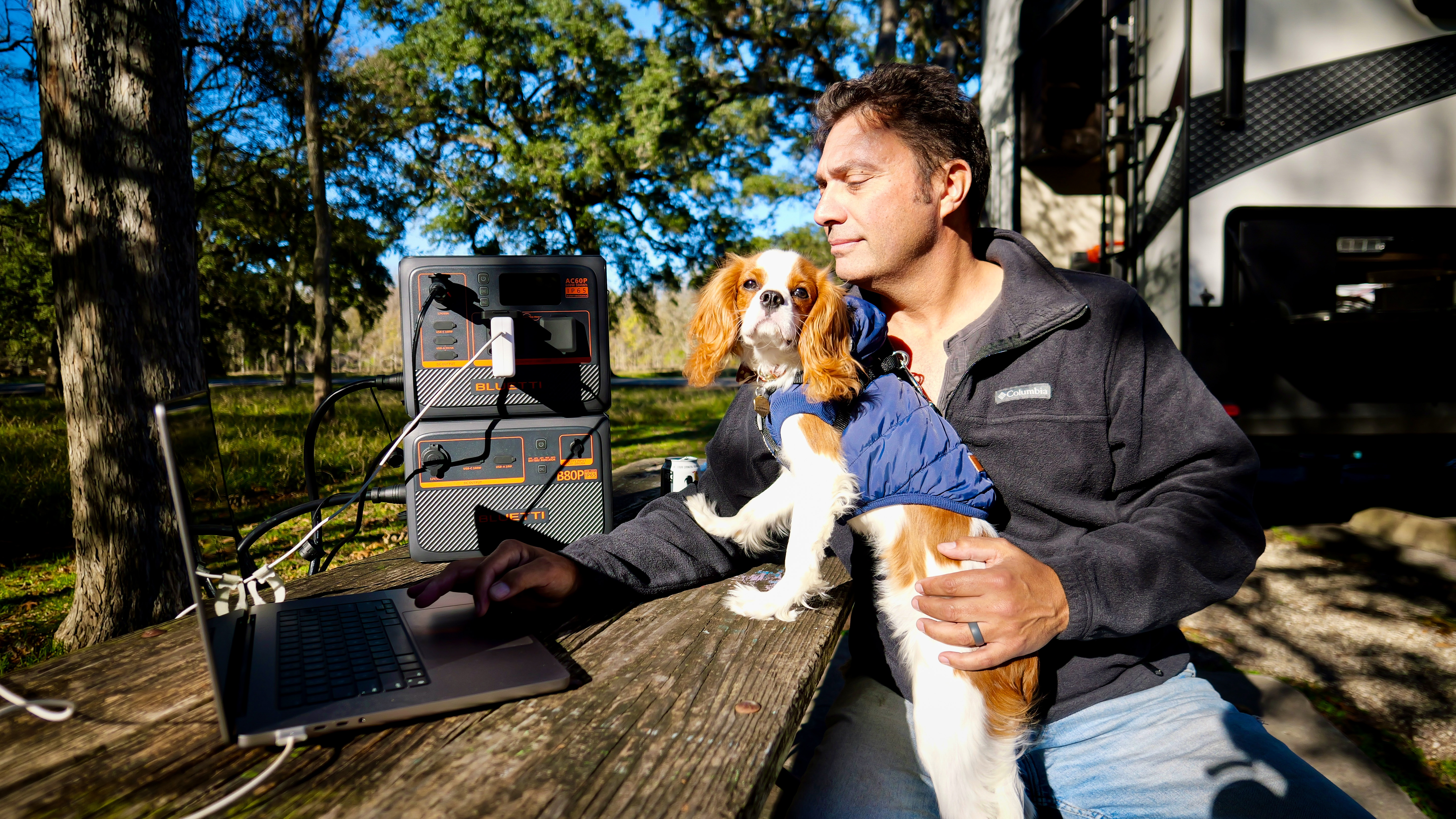 Mężczyzna pracuje na laptopie przy stole piknikowym, z psem i stacją zasilania.