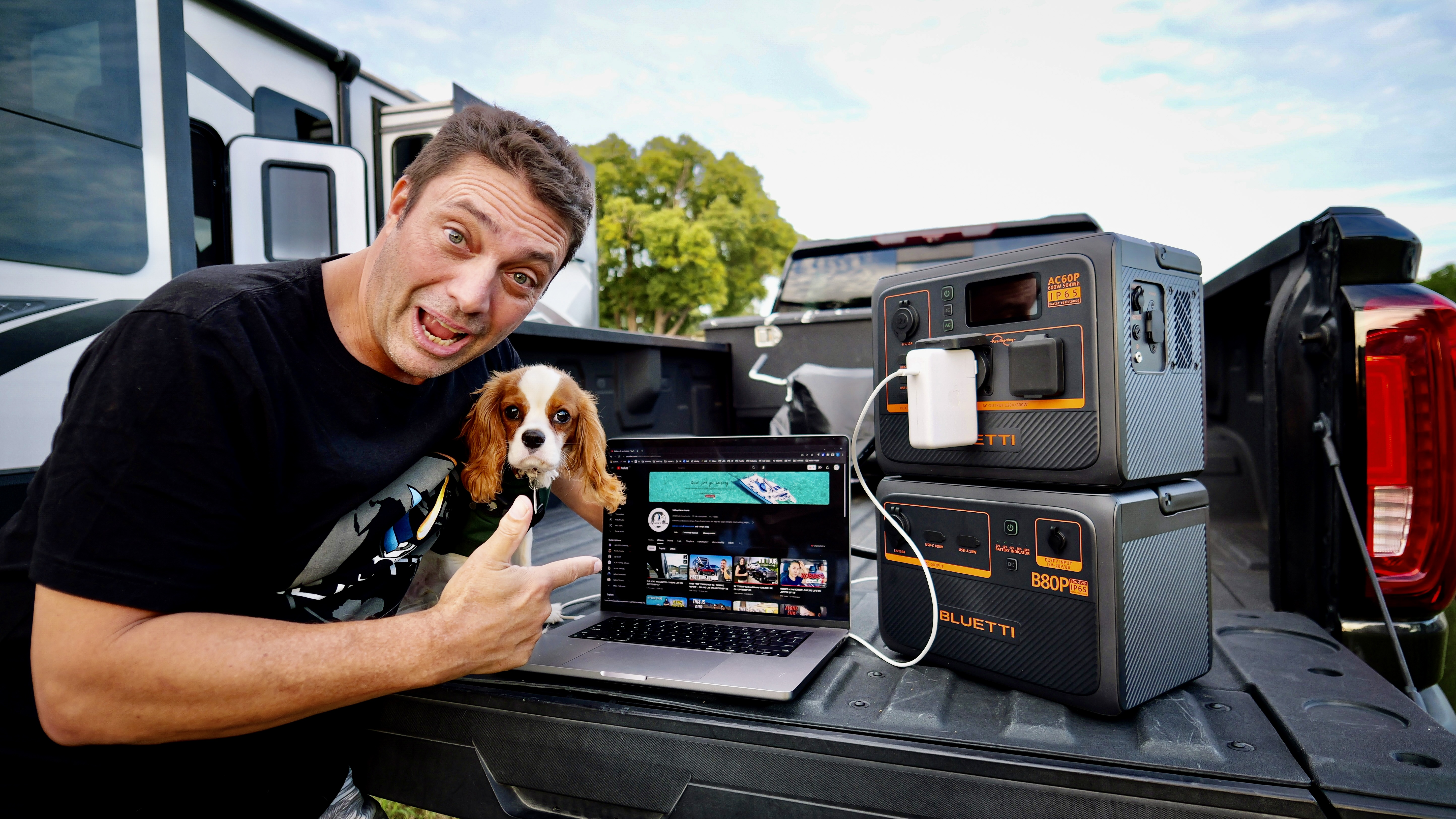 Mężczyzna i pies w pobliżu pojazdu, używający laptopa i stacji zasilania na zewnątrz.