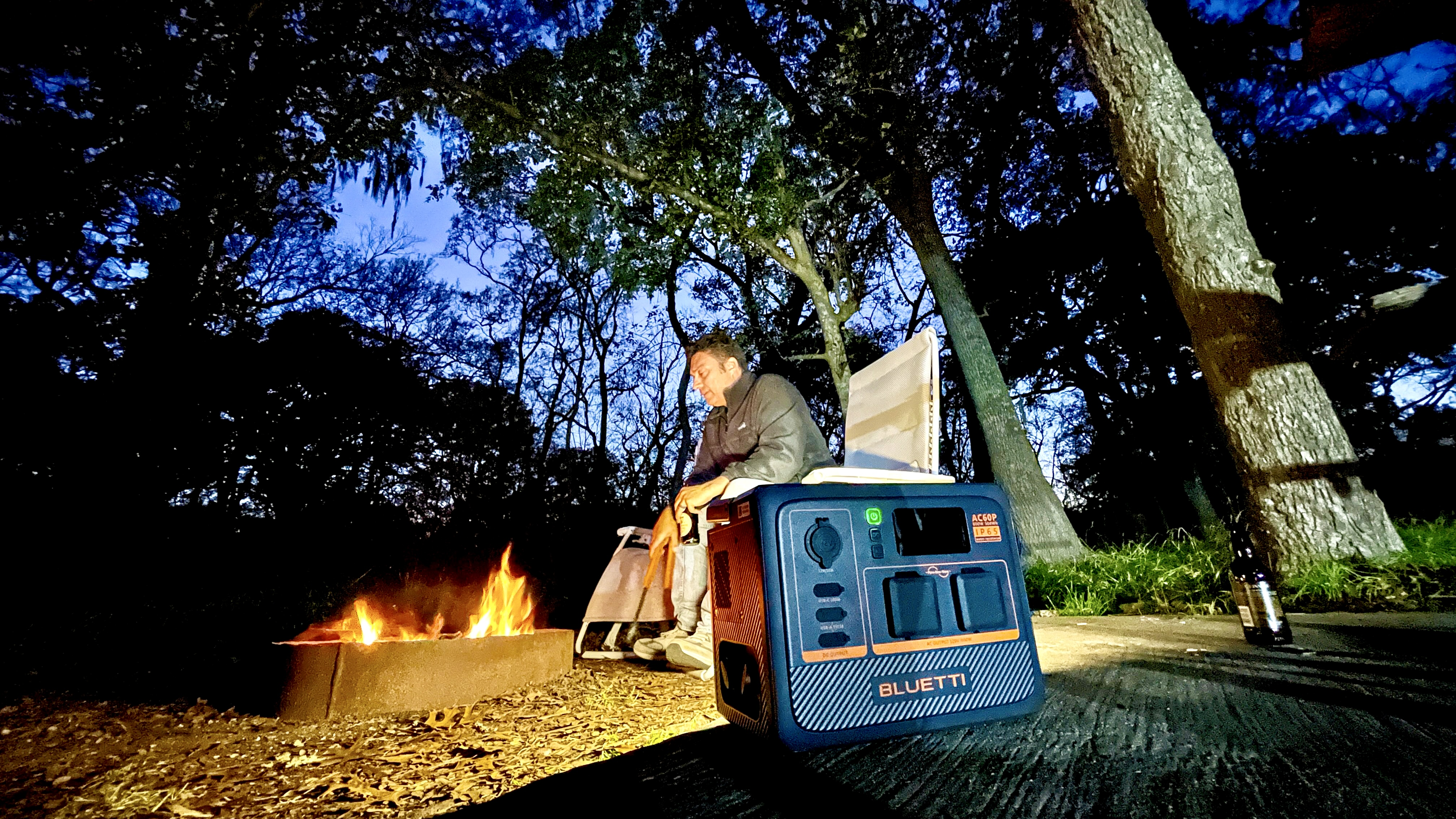 Osoba siedzi przy ognisku, z elektrownią Bluetti i drzewami w tle.