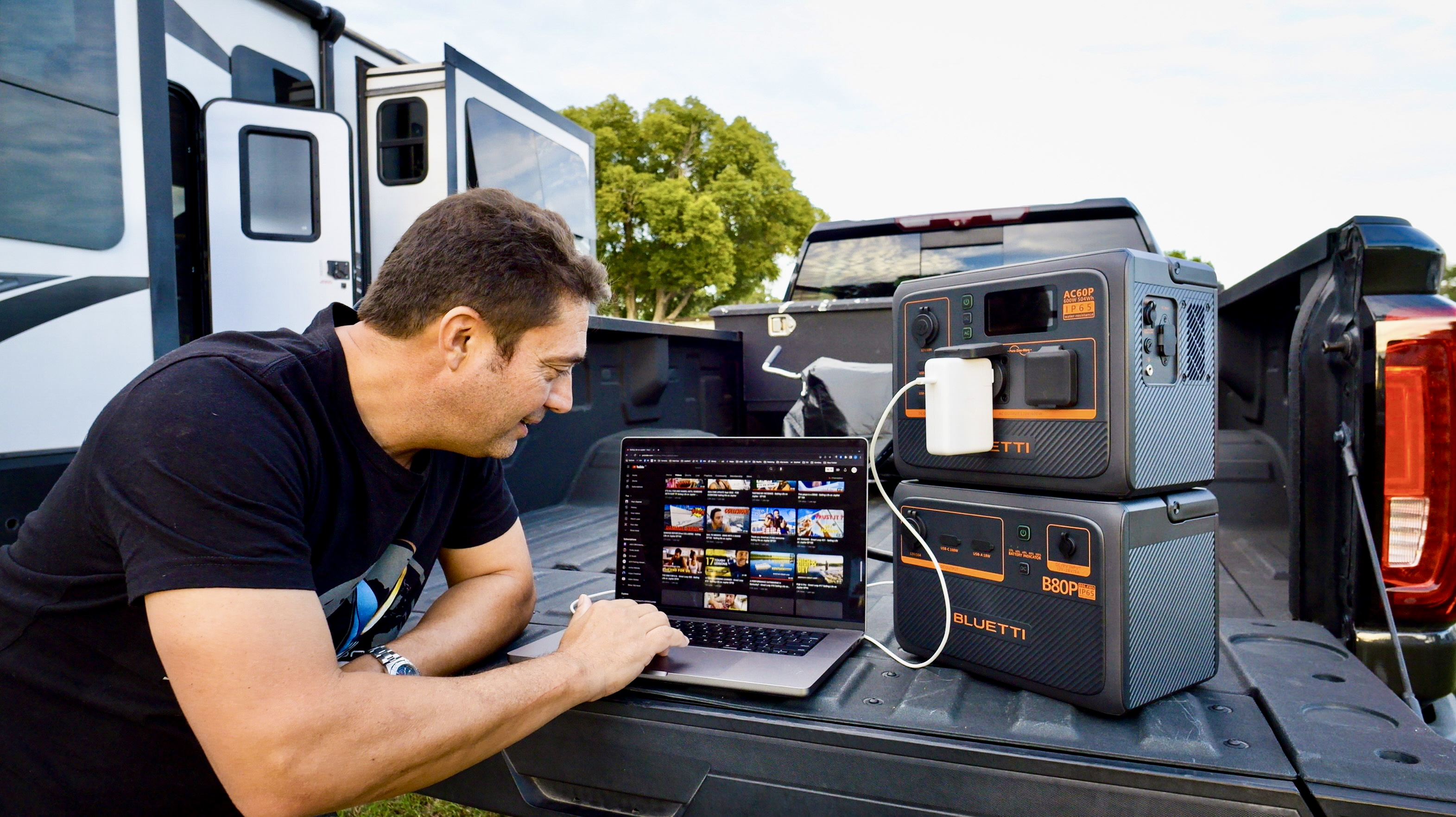 Mężczyzna pracujący na laptopie na zewnątrz. Laptop jest podłączony do elektrowni, samochód zaparkowany w tle.